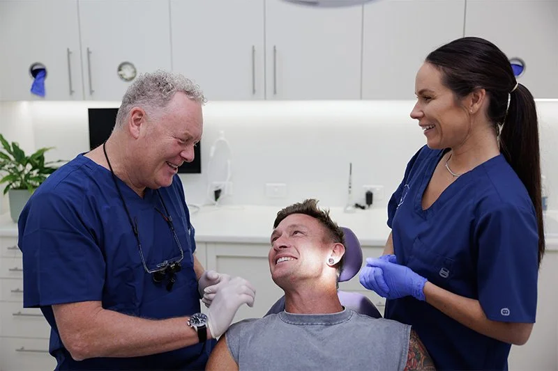 A man sitting in a dental chair smiling with dental professionals, a male dentist and a female dental assistant, attending to him in a dental office.