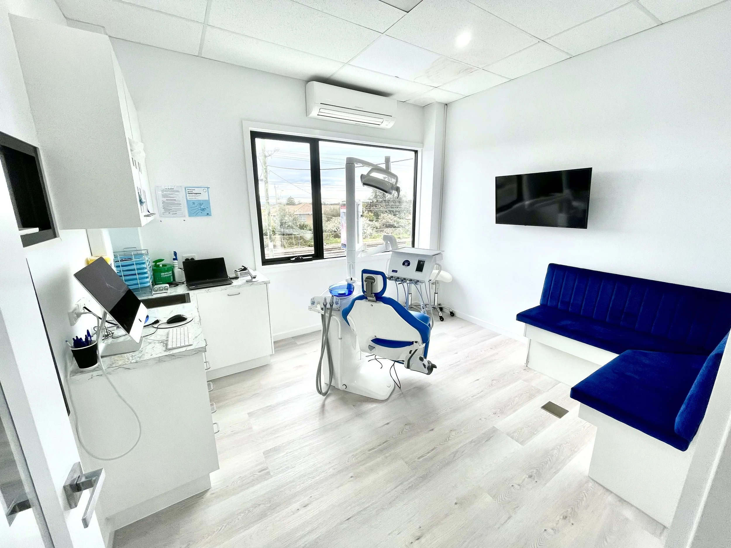 A clean dental office with a dental chair near a window, a computer on a white counter, a blue bench, and dental equipment in the room.