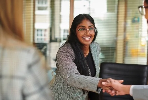 A woman smiling and shaking hands with a man in a professional setting, with another person blurred in the foreground.