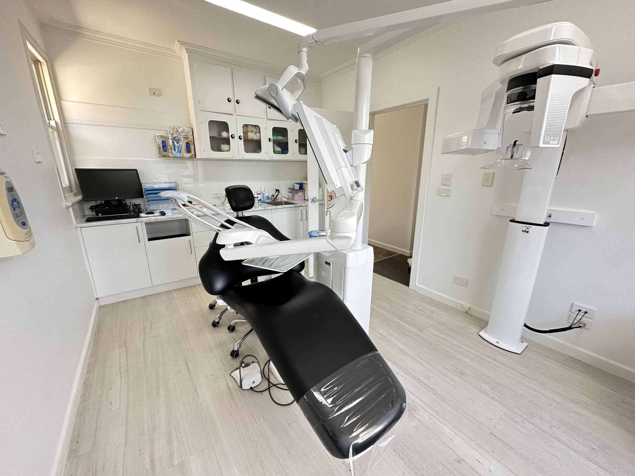 Dental examination room with dental chair, X-ray machine, computer, and white cabinets.