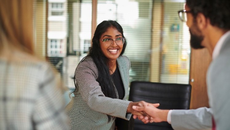 A woman with glasses and a gray blazer shaking hands with a man in a business setting, smiling.