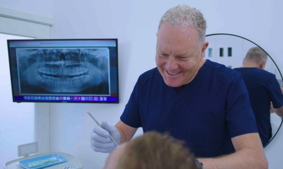 A dentist in navy scrubs smiling and holding a dental tool while examining a patient in a dental office with a digital x-ray of teeth on a monitor in the background.