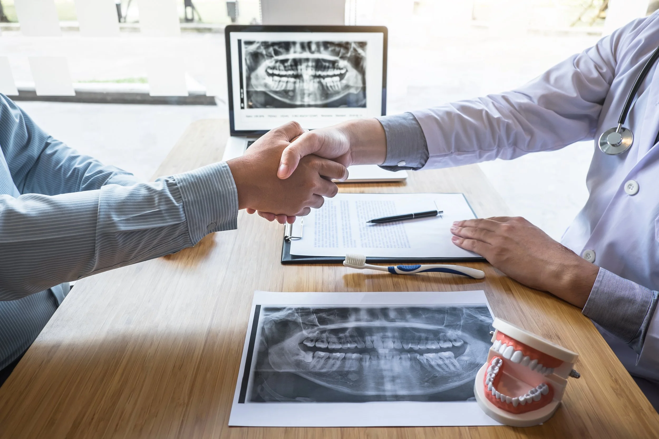 A dentist and a patient shaking hands at a dental appointment. On the table are dental X-rays, a model of teeth, and dental tools, with a computer displaying a panoramic dental X-ray in the background.