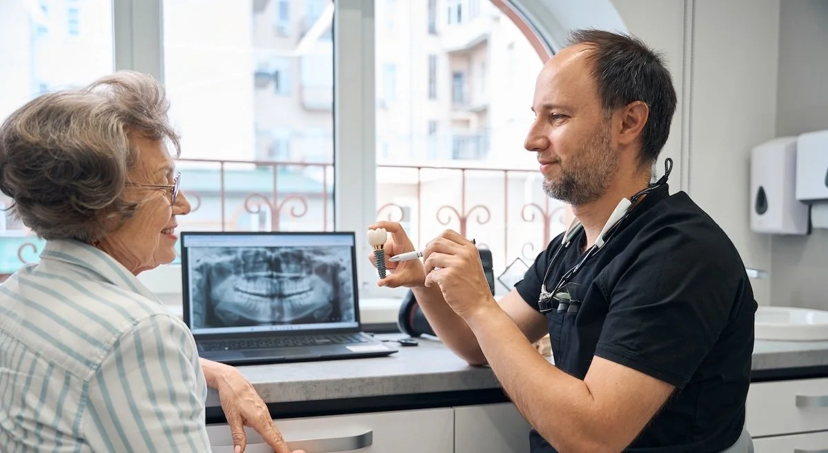 Dentist showing a dental implant model to an elderly patient in a dental clinic, with an x-ray on the laptop screen.