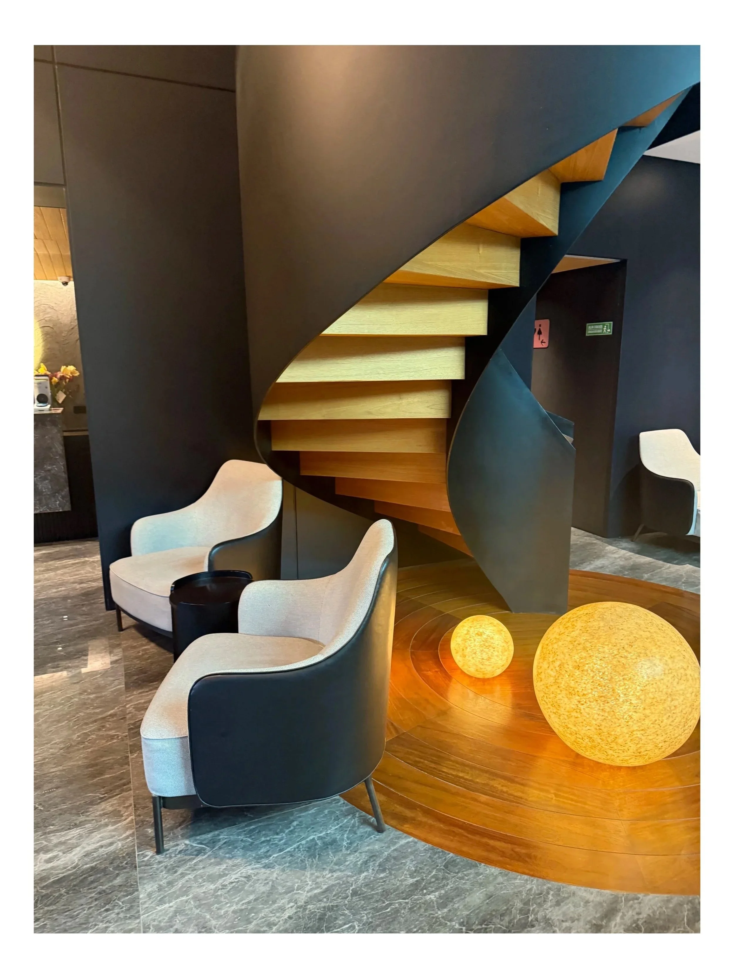 Modern lobby with black spiral staircase, beige armchairs, and glowing spherical lamps on wooden floor.