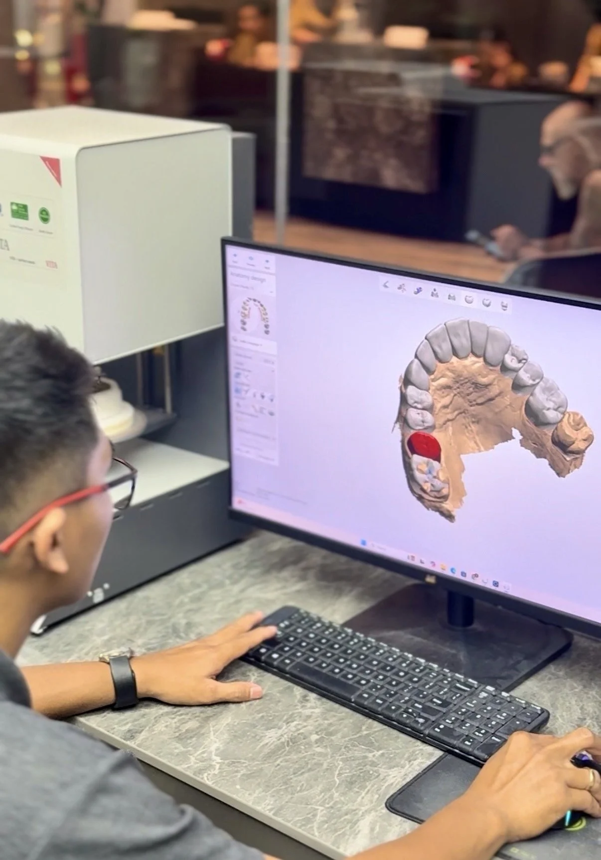 A person working on a computer designing a 3D model of a human jaw with teeth, wearing glasses and a watch, in an office or clinic setting.