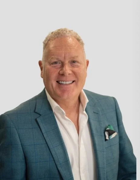 A smiling man with curly blonde hair, wearing a blue checked suit jacket, white shirt, and a pocket square, standing against a plain background.