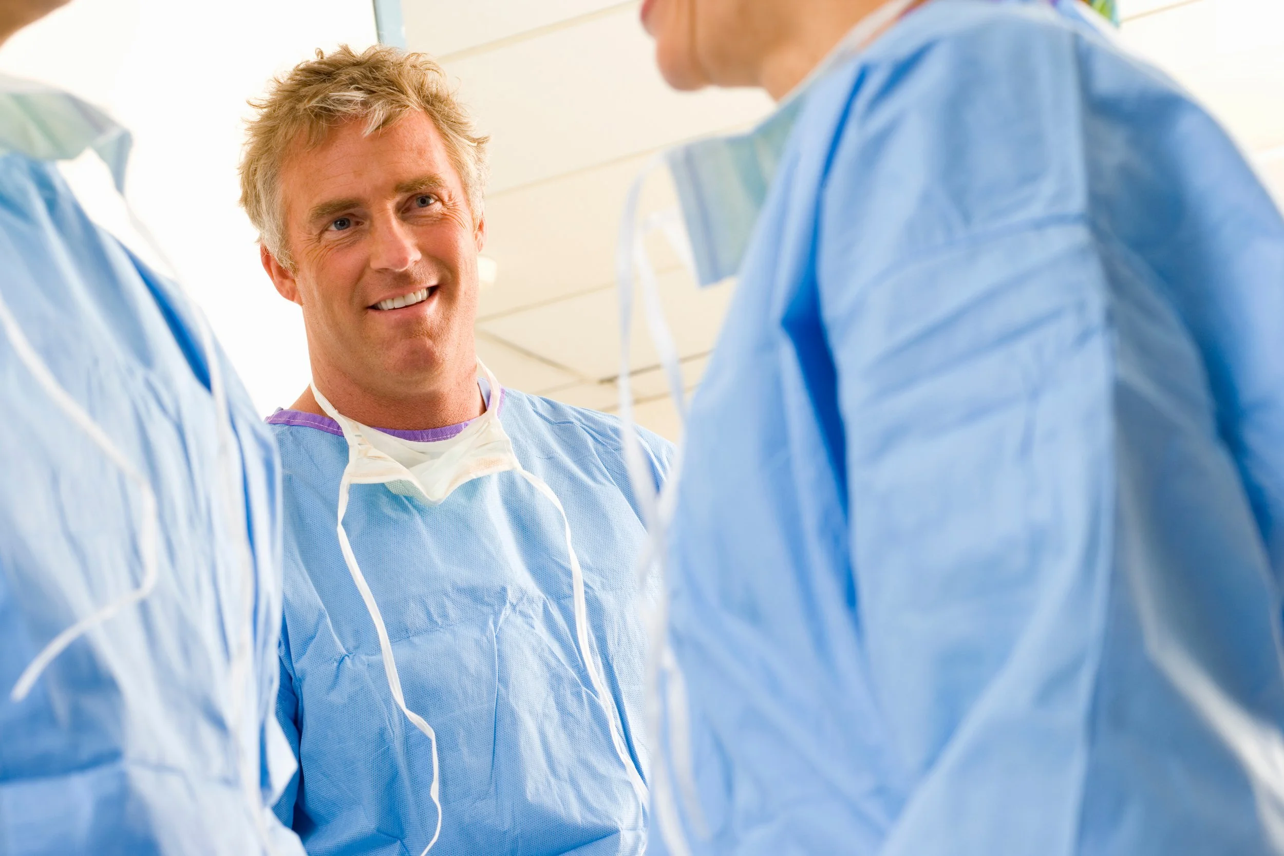 A smiling male doctor or nurse in blue scrubs talking to two other medical professionals in an examination or hospital setting.