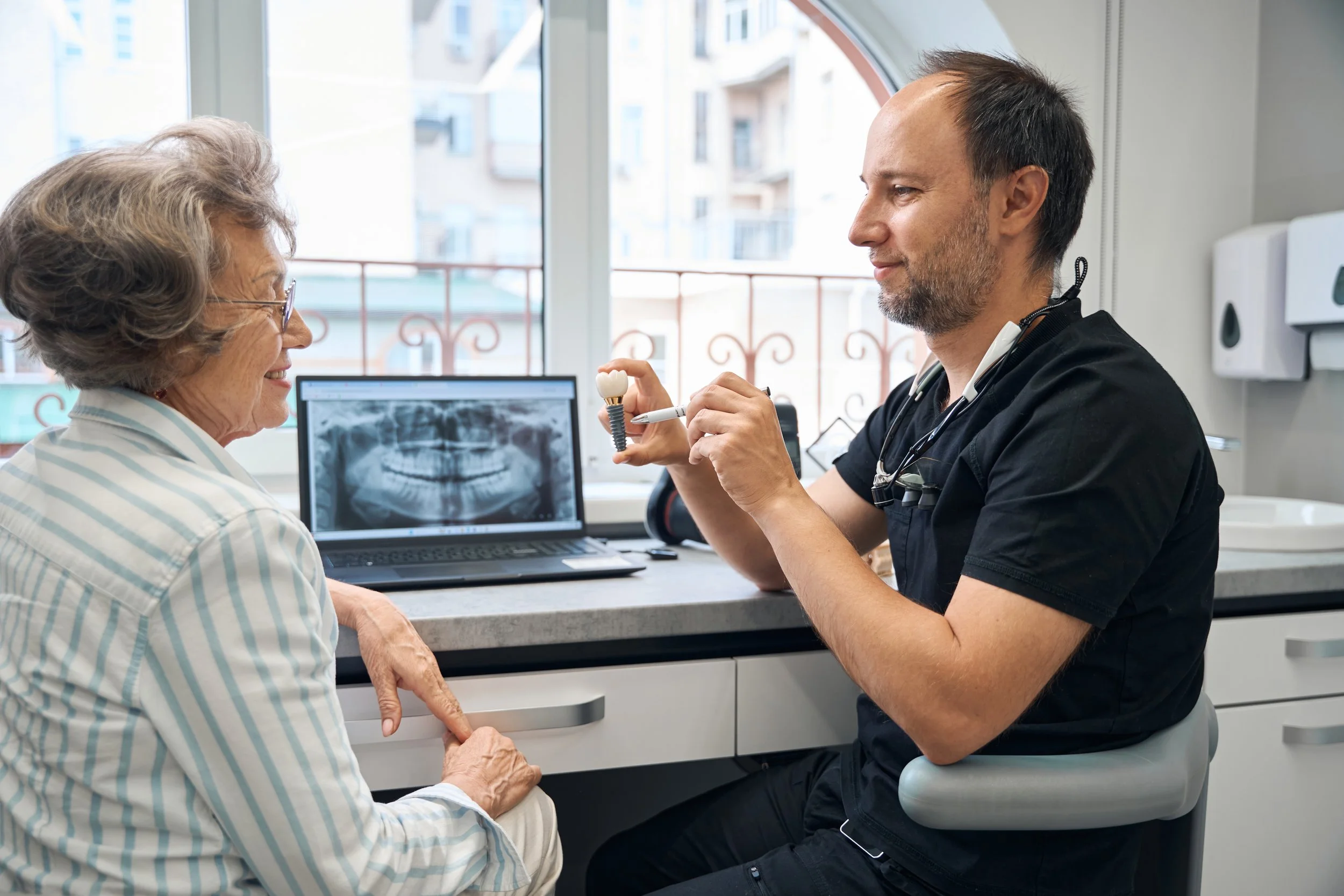 A dentist showing a model of a tooth and dental implant to an elderly woman at a dental office, with an X-ray displayed on a laptop in the background.