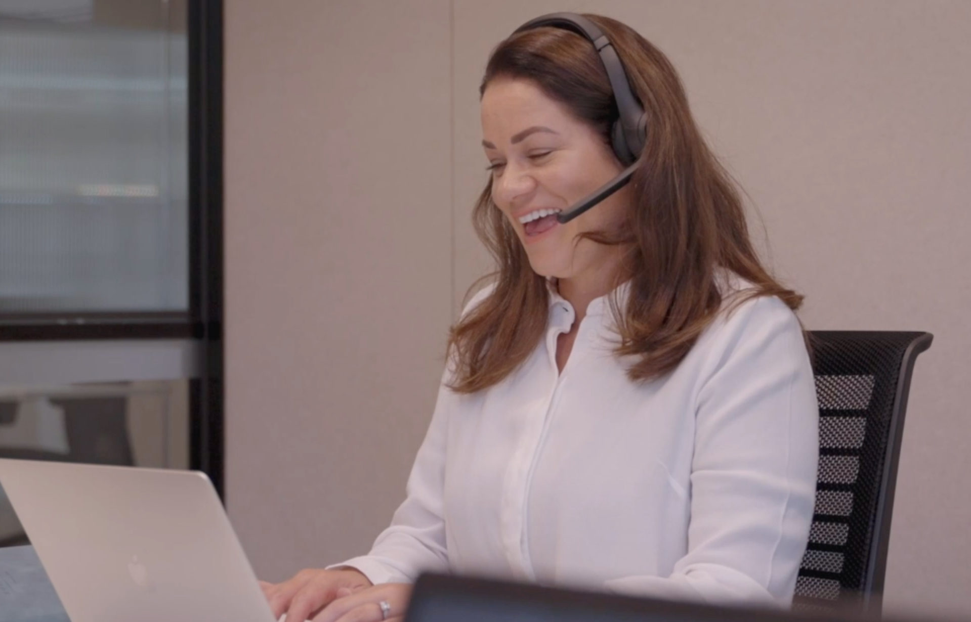 A woman with brown hair wearing a headset and white shirt, smiling while working on a laptop in an office setting.