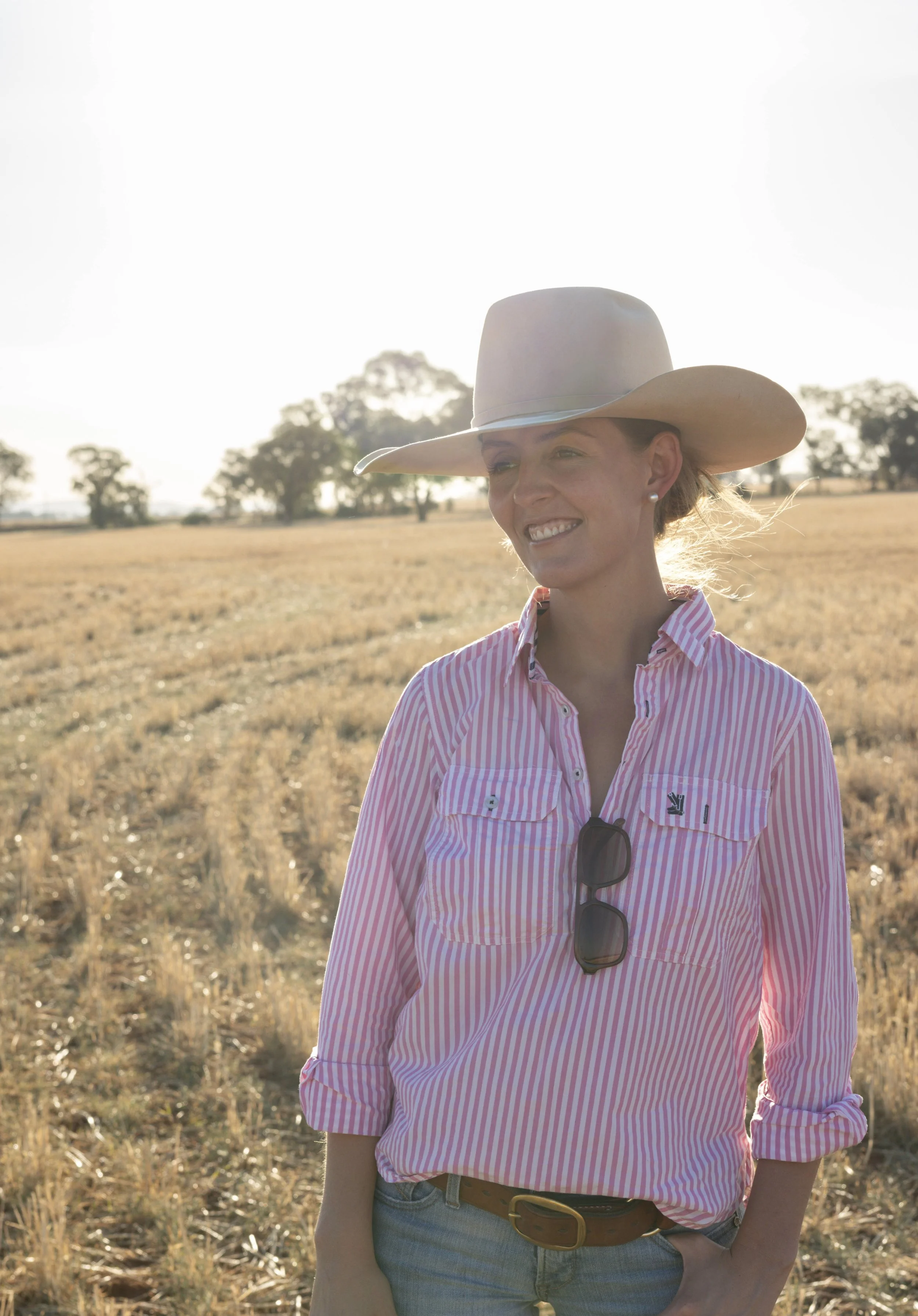 Katie Doyle from Mud Media, smiling in a field during sunset, wearing a cowboy hat, pink striped shirt, sunglasses hanging from her shirt, and jeans.