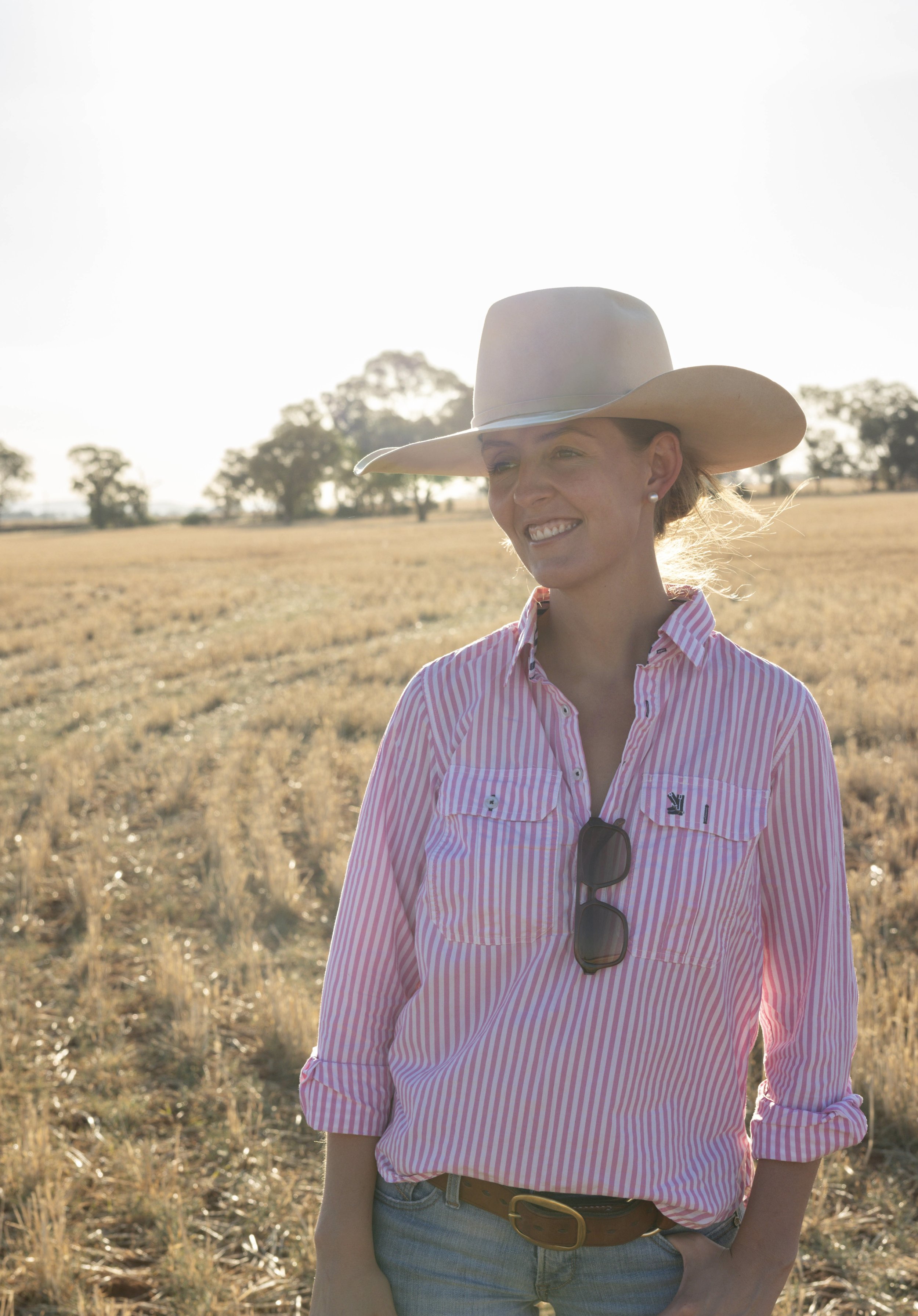 Katie Doyle from Mud Media, smiling in a field during sunset, wearing a cowboy hat, pink striped shirt, sunglasses hanging from her shirt, and jeans.