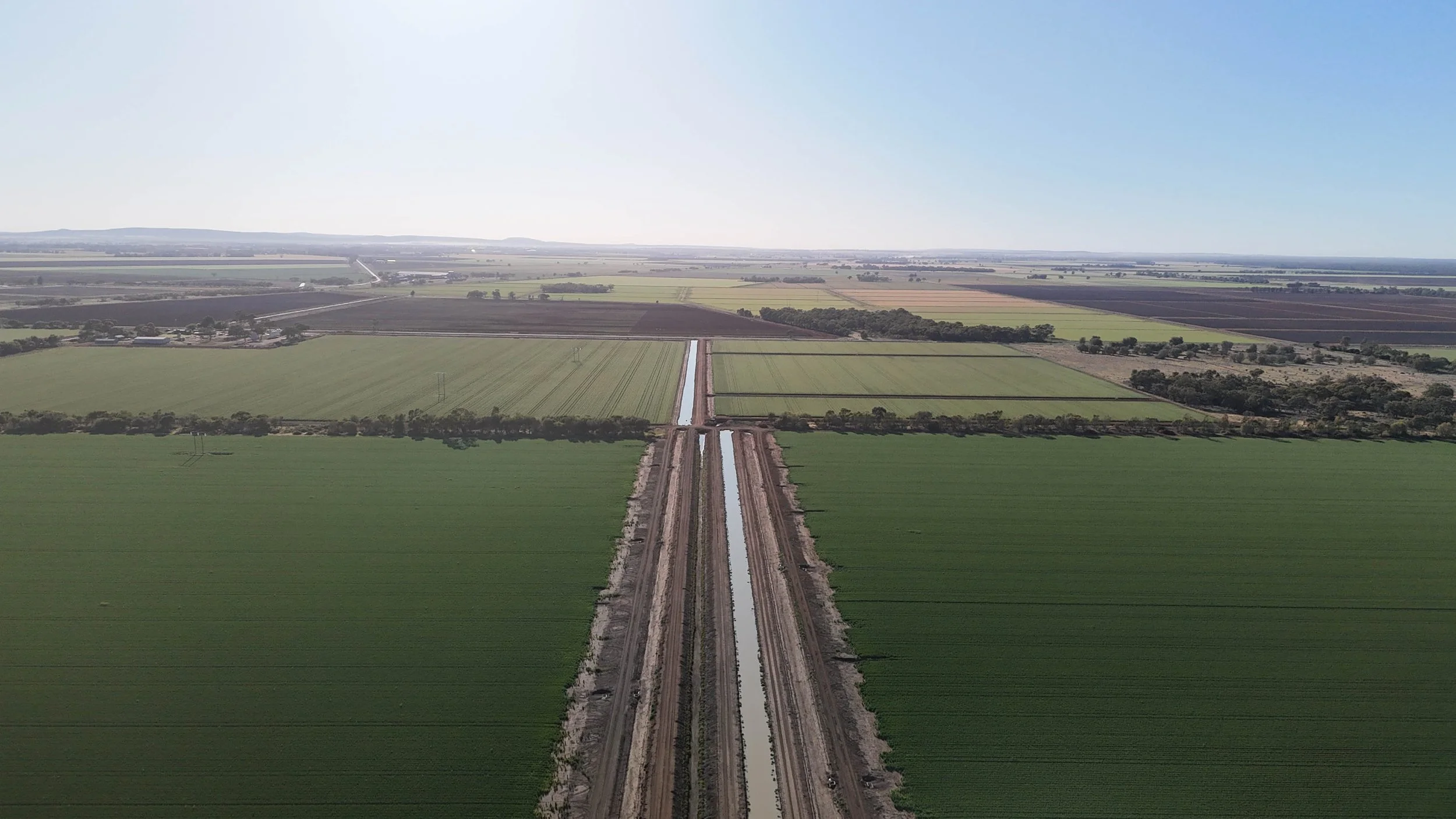 Aerial view of farmland with a new irrigation canal and dirt roads, surrounded by green crop fields under a clear sky.