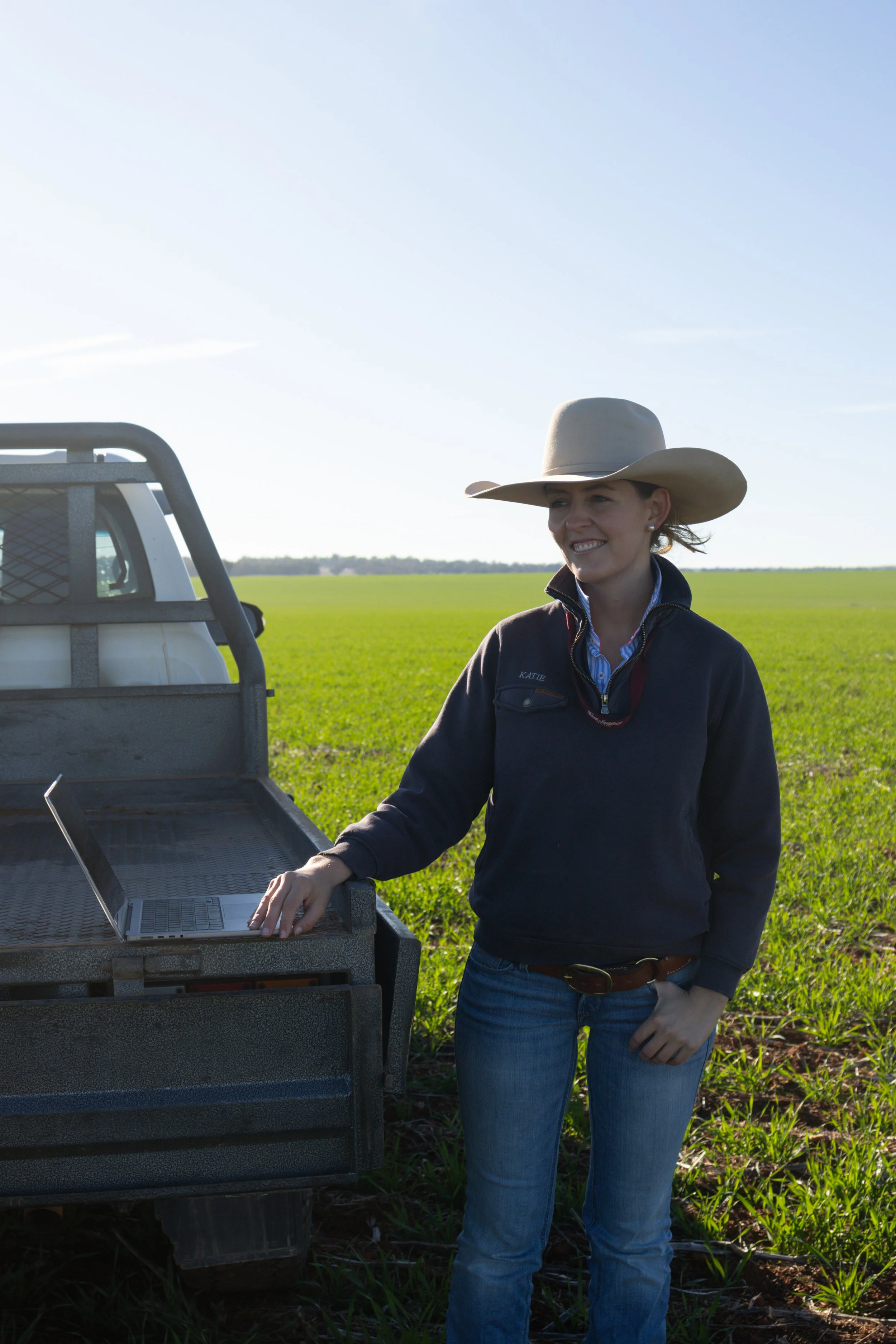 Katie Doyle, standing outdoors in a green field, wearing a wide-brimmed hat, a dark jacket, and jeans, with a laptop on the back of a pickup truck.