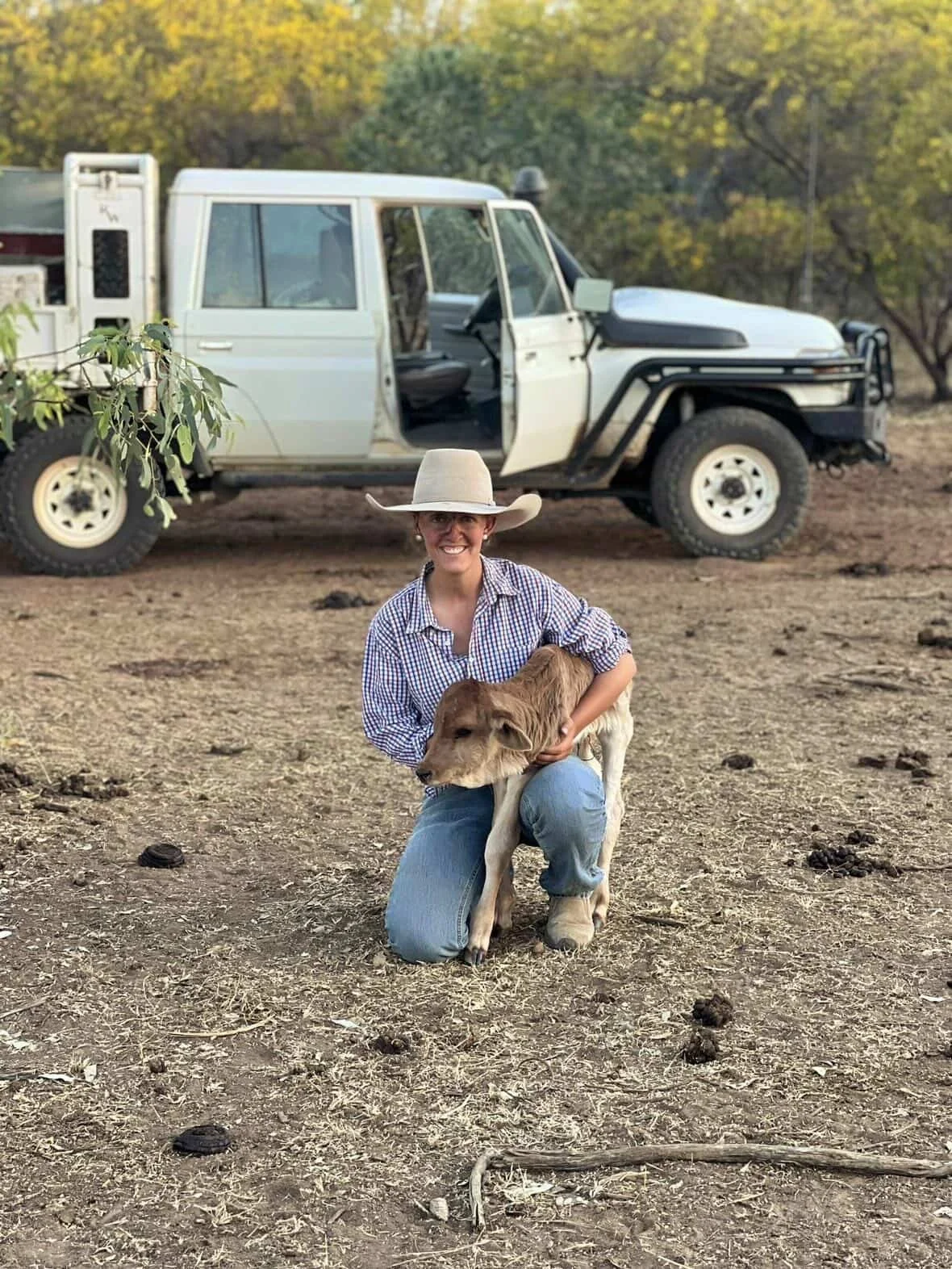 A woman wearing a large wide-brimmed hat and a checkered shirt kneeling on dirt ground, holding a young calf. A white off-road vehicle with an open door is parked behind them. There are trees with green and yellow leaves in the background, indicating a fall setting.