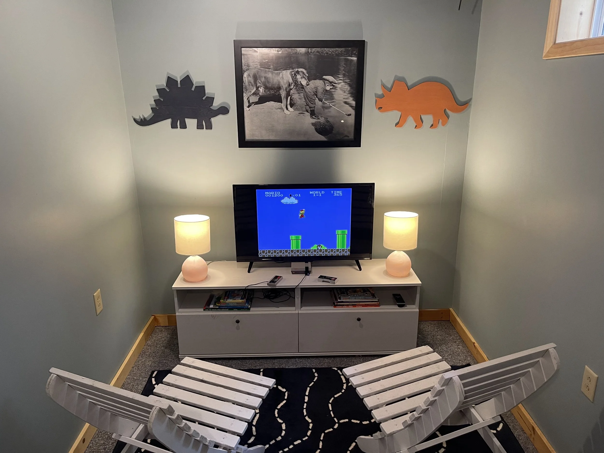 Cozy basement entertainment area with a small television showing a classic video game, two white chairs in front, two matching lamps on either side of the TV, wall decorations of a black and white photo of a dog and a child by the water, and wooden d