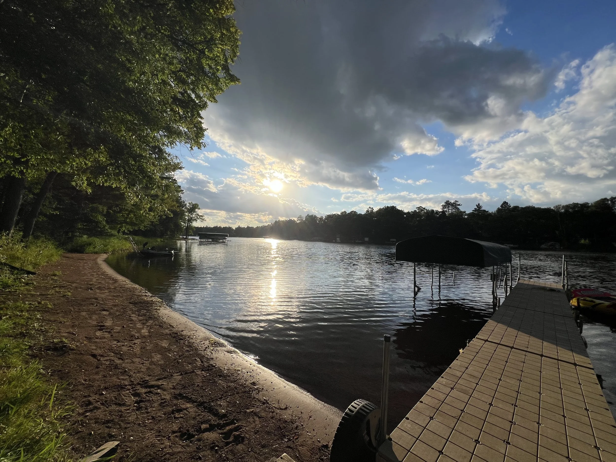 Sunset over a calm lake with a dock extending into the water, surrounded by trees under a partly cloudy sky.