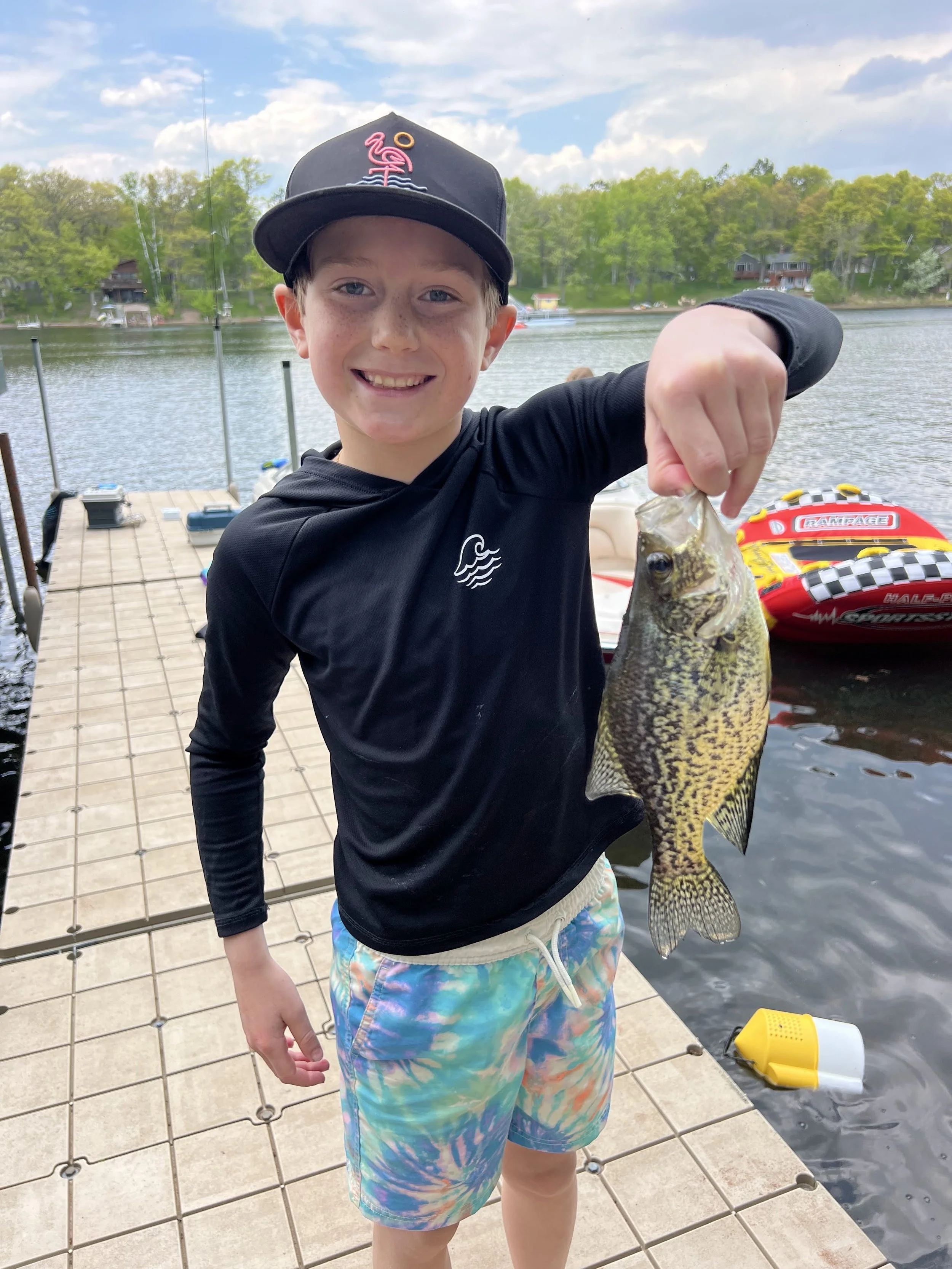 A young boy smiling and holding a largemouth bass he caught, standing on a dock with water and trees in the background, wearing a black cap and a black long-sleeve shirt.