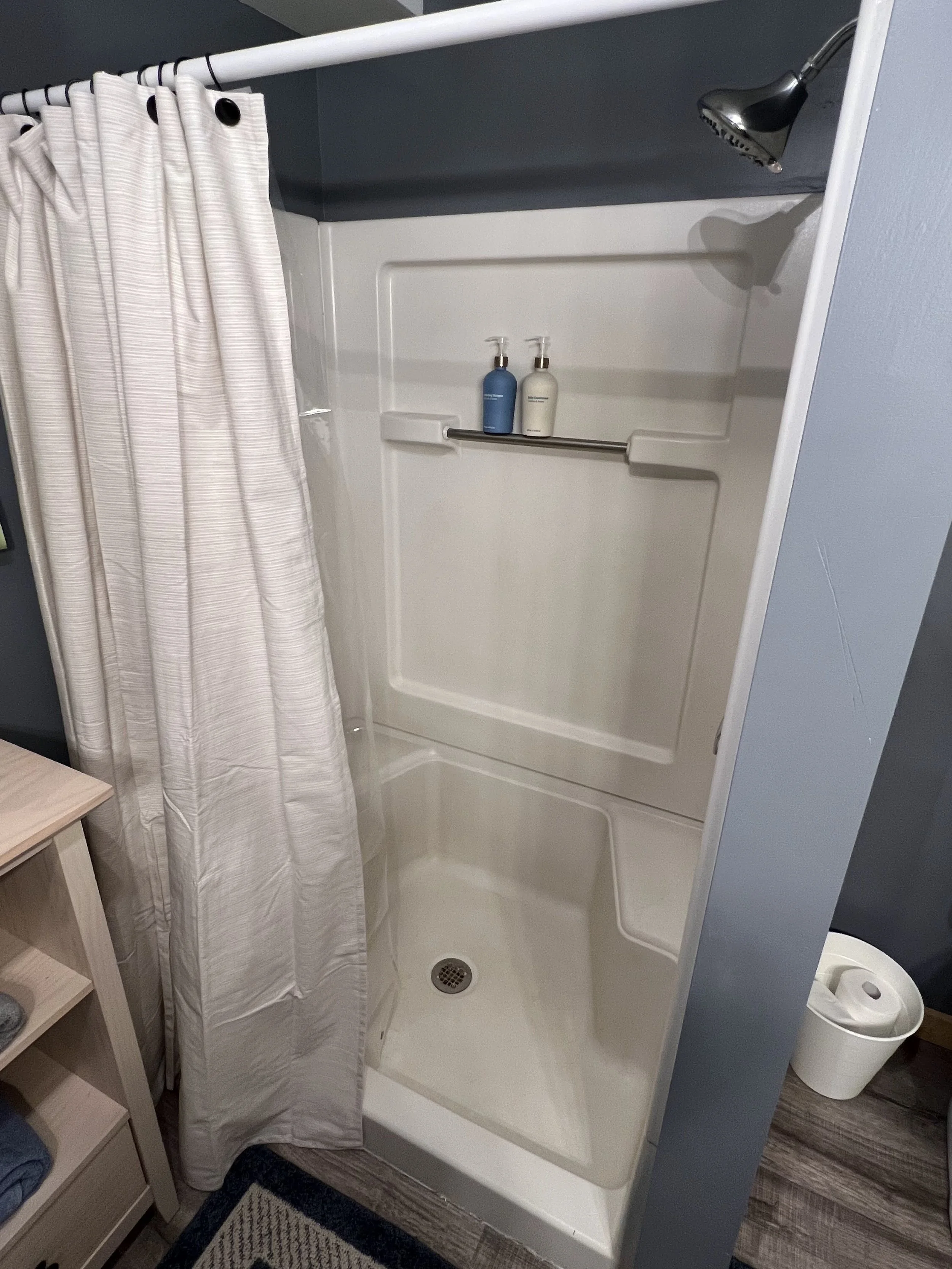 Small shower with white walls, a beige curtain, two bottles of shampoo on a metal bar, and a round drain on the floor.