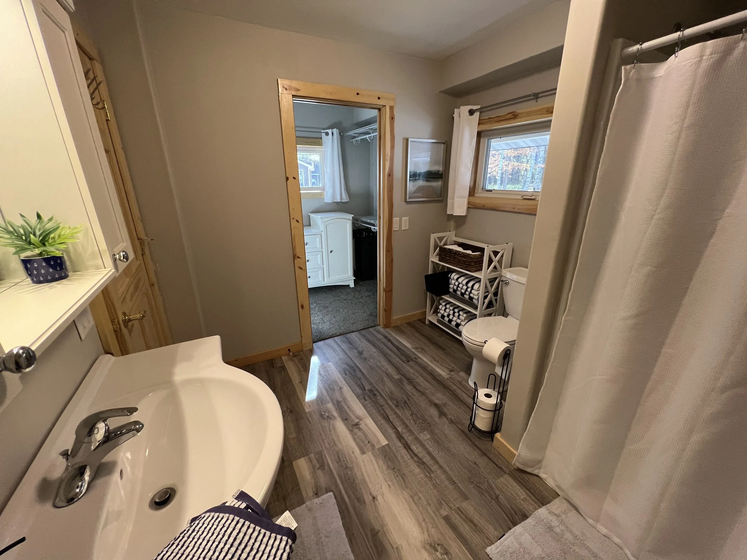 Bathroom with wooden trim, featuring a white sink with a striped towel, toilet with toilet paper, small white shelving unit with black and white towels, window with white curtains, and a doorway leading to a laundry area with a white cabinet and wind
