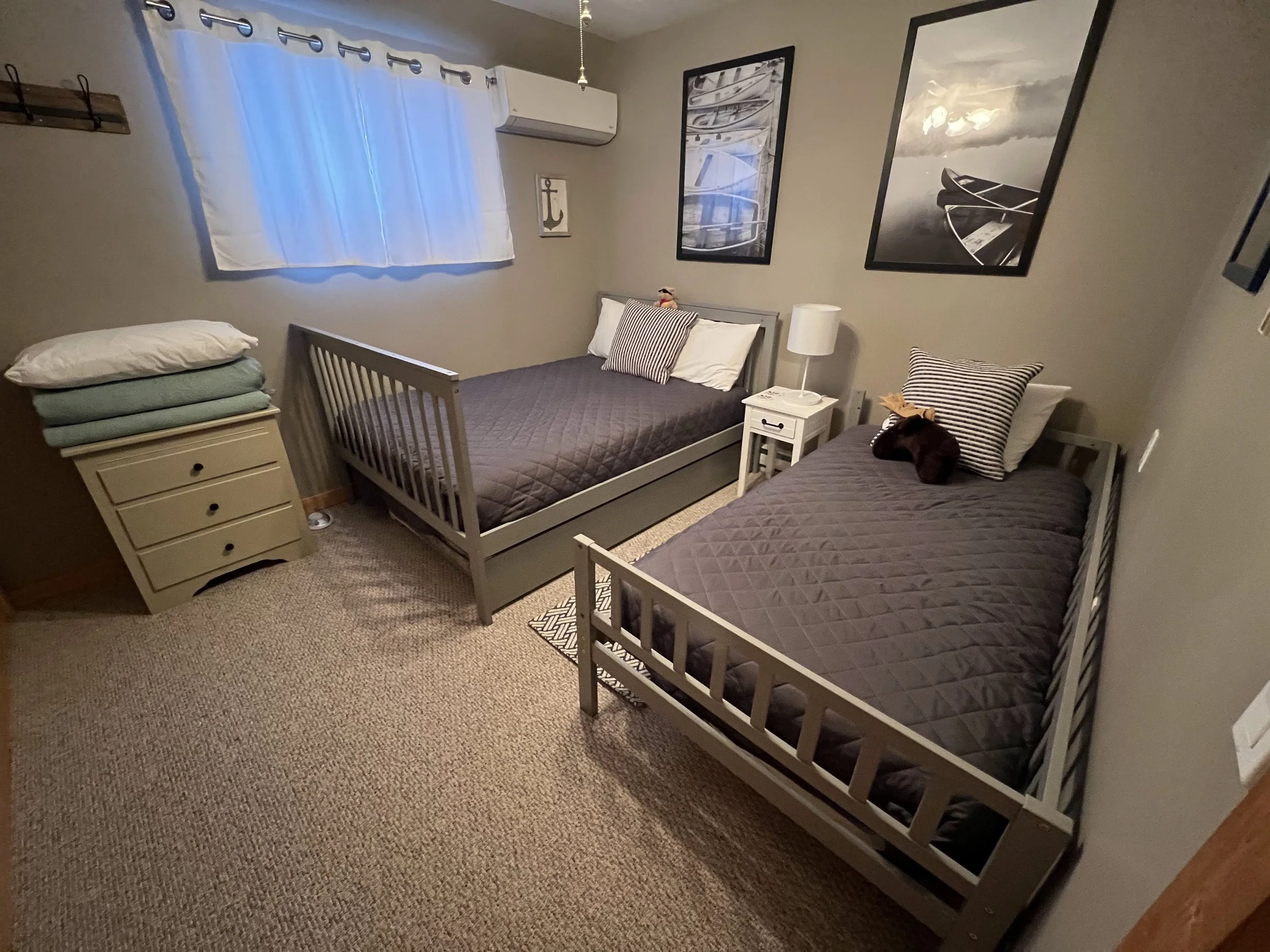 A bedroom with two twin beds with gray frames, one with pillows and a stuffed animal, the other with pillows and a black stuffed bear. There is a window with white curtains, a small white nightstand with a lamp, framed nautical-themed artwork on the 