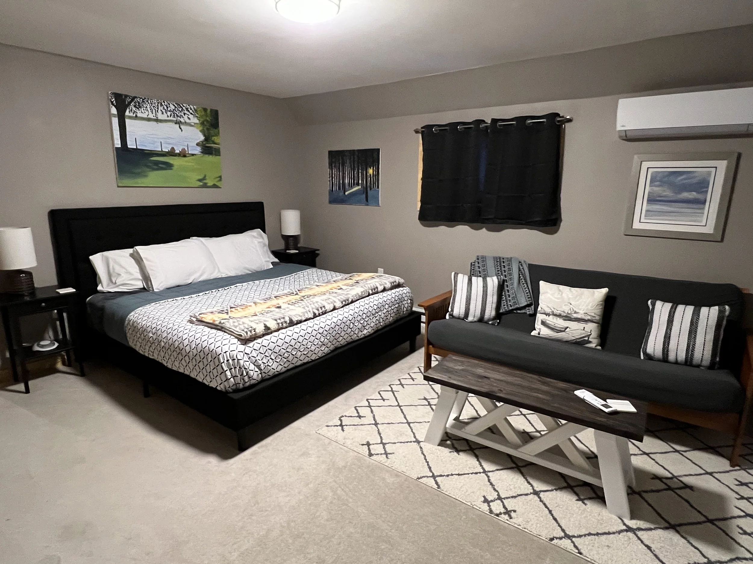 A bedroom with a black bed frame, white pillows, patterned bedspread, black side tables with lamps, a black sofa with striped and patterned pillows, a wooden coffee table, a white area rug, framed artwork, and a window with black curtains.