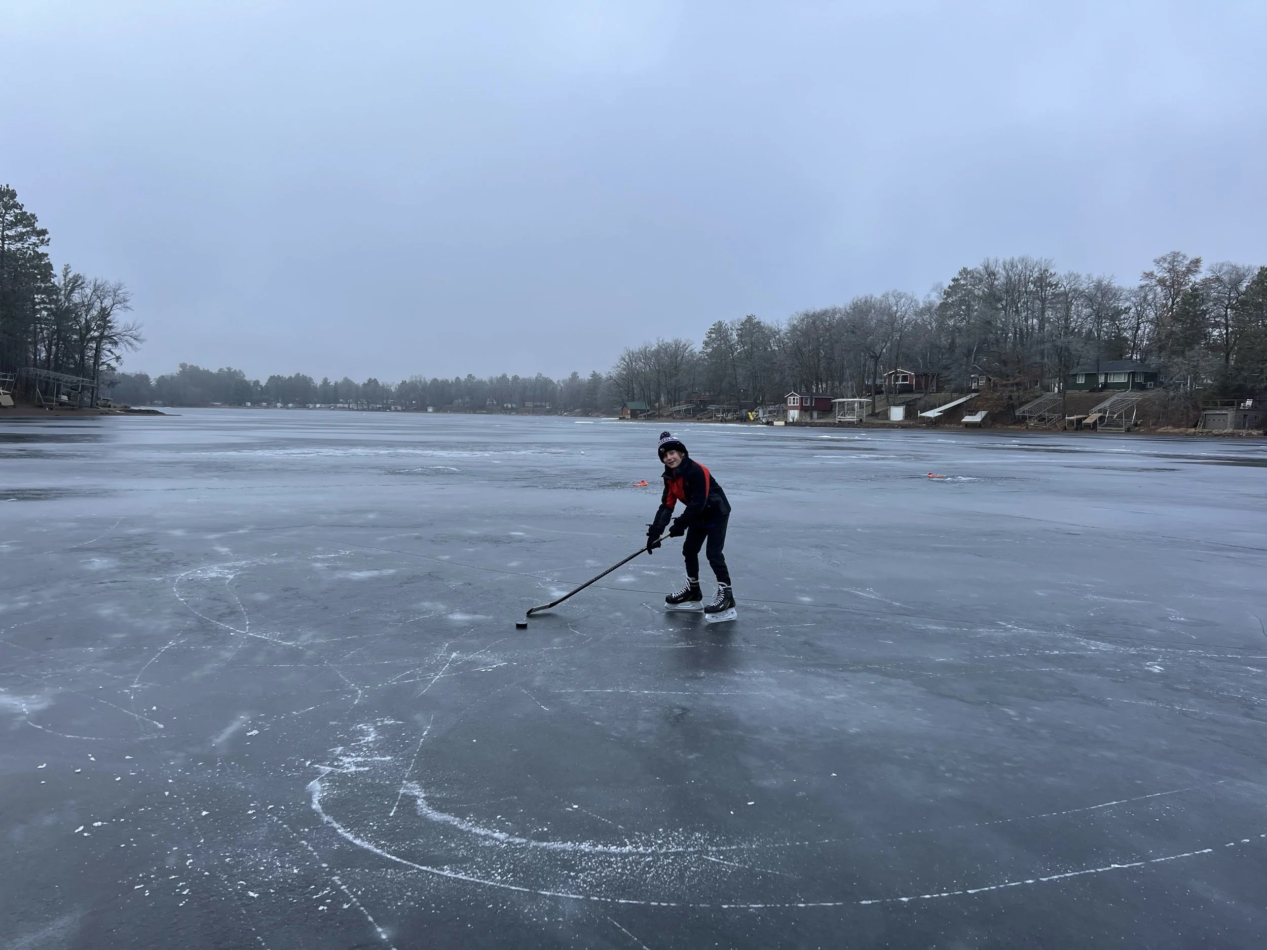 Person skating with a hockey stick on a frozen lake, with houses and trees in the background on a cloudy day.