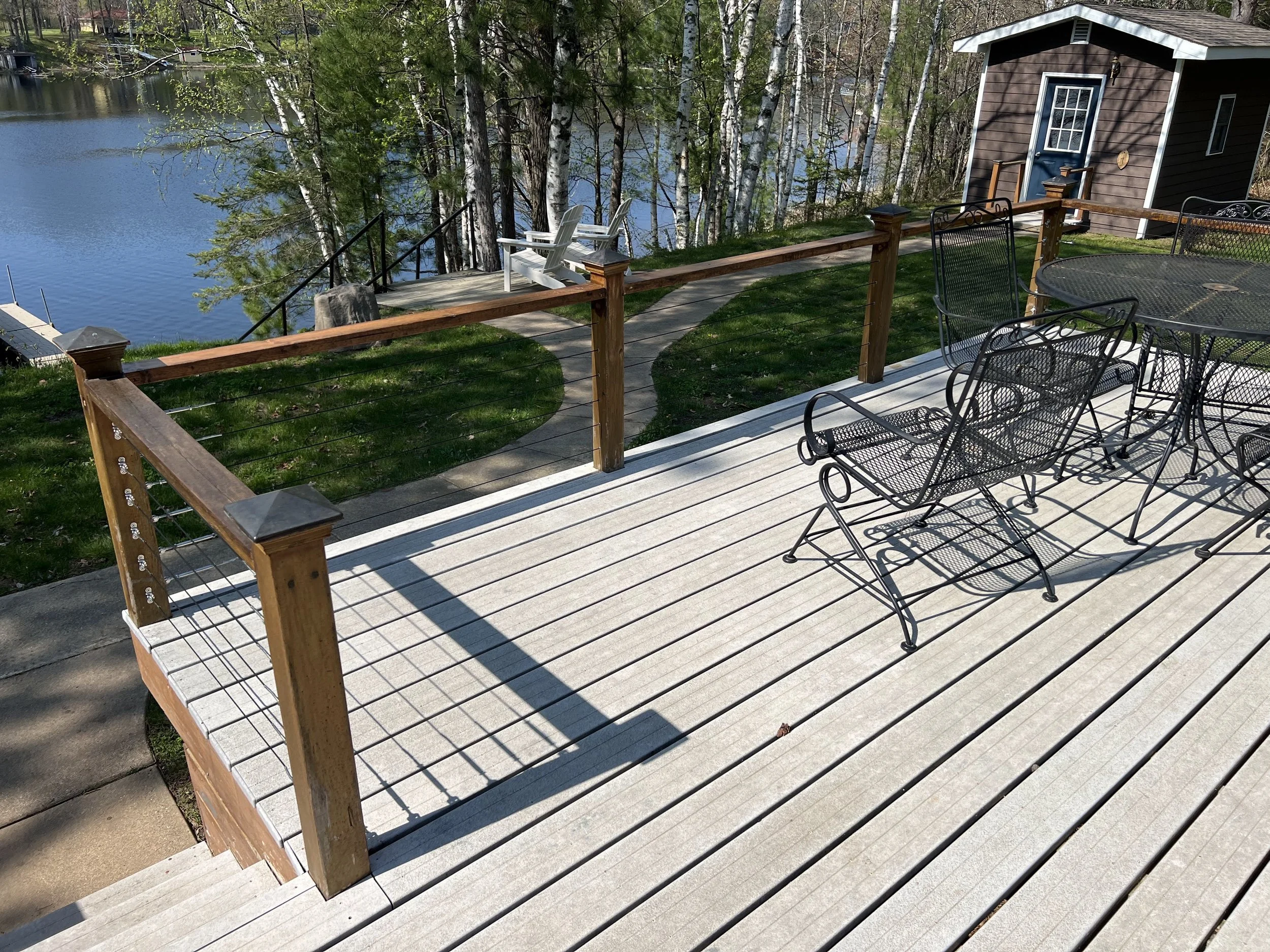 Backyard deck with black metal patio furniture, overlooking a pond and trees, with a small shed and two white Adirondack chairs near the water.