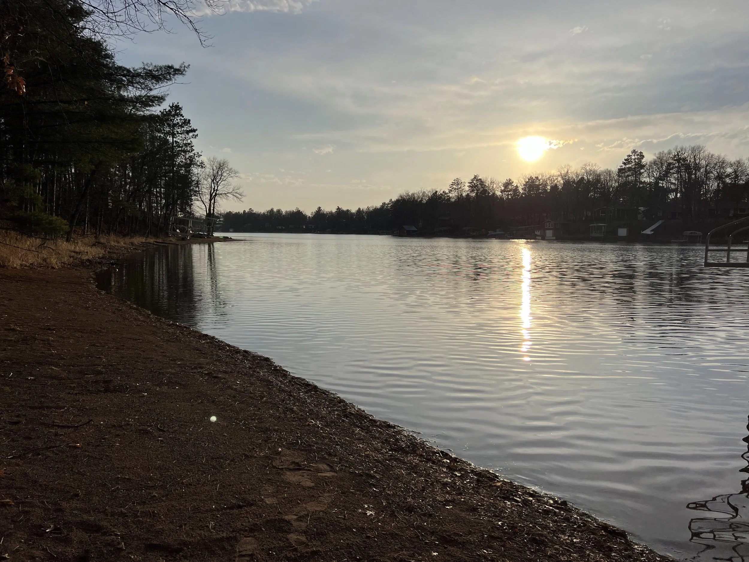 Sun setting over a calm river, with trees along the shoreline and houses in the background.