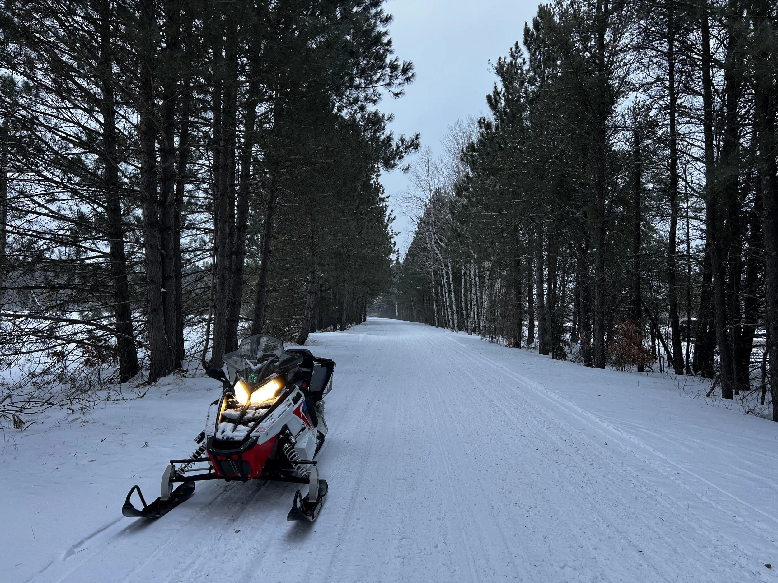 A snow-covered trail through a forest with pine trees on both sides and a snowmobile parked on the left side of the trail.