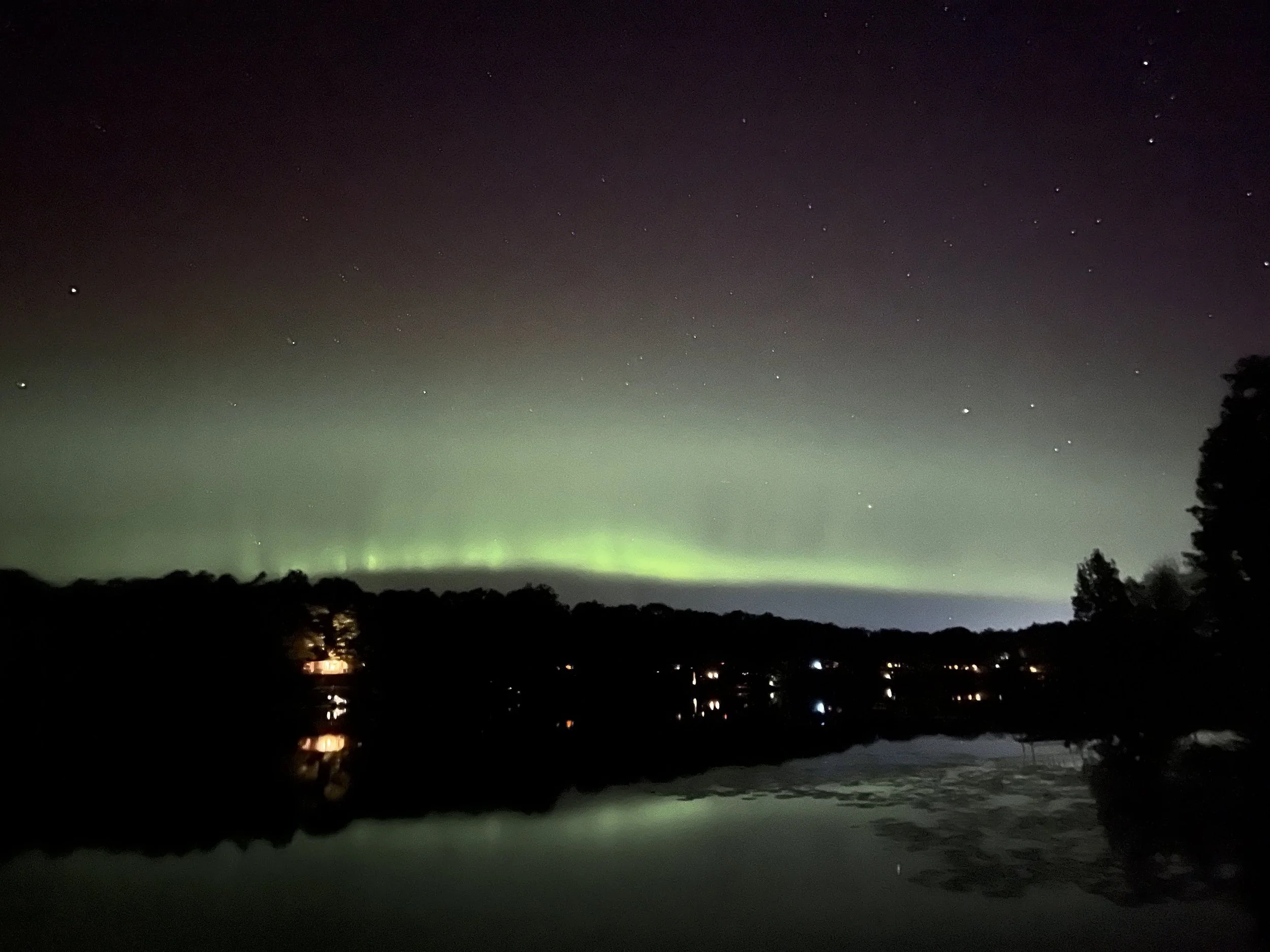 Night sky with the Northern Lights over a lake with reflections, silhouette of trees and scattered lights from houses.
