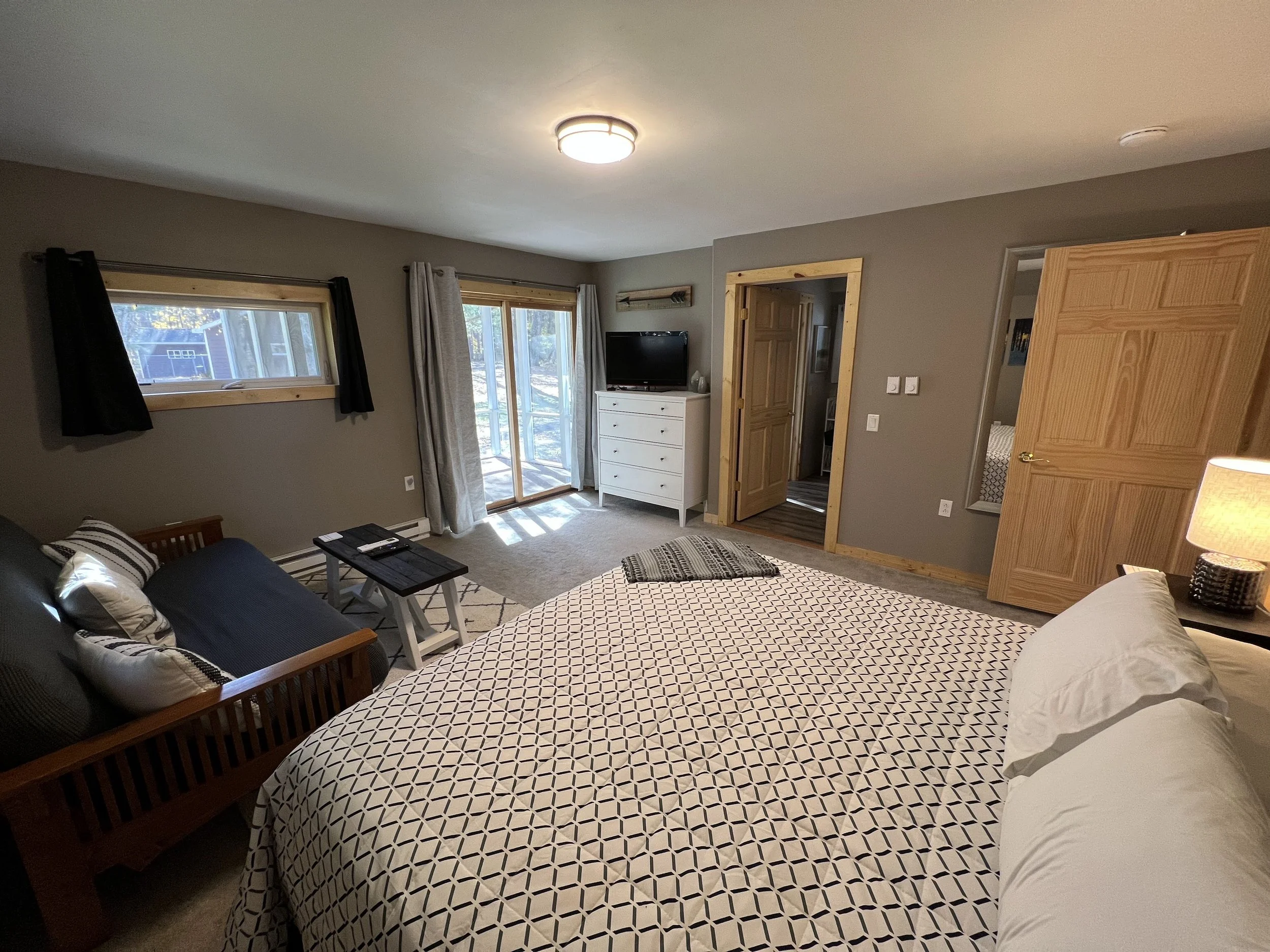 A cozy bedroom with a queen-sized bed, a small black sofa with pillows, a white dresser with a television, a glass door leading to an outdoor area, and windows with black and beige curtains.