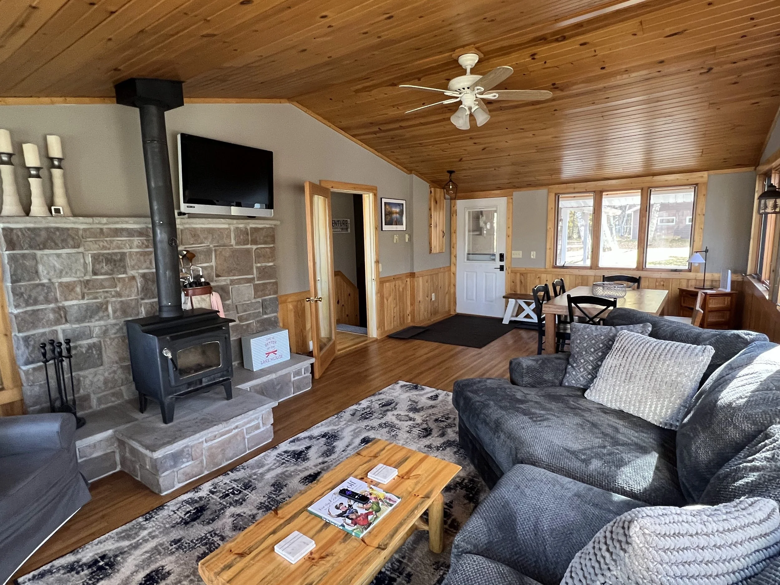 Living room with gray sofa, wooden coffee table, stone fireplace with a chimney, wall-mounted TV, light wood paneled ceiling, ceiling fan, large windows, dining table with chairs, wooden accents, and hardwood floors.