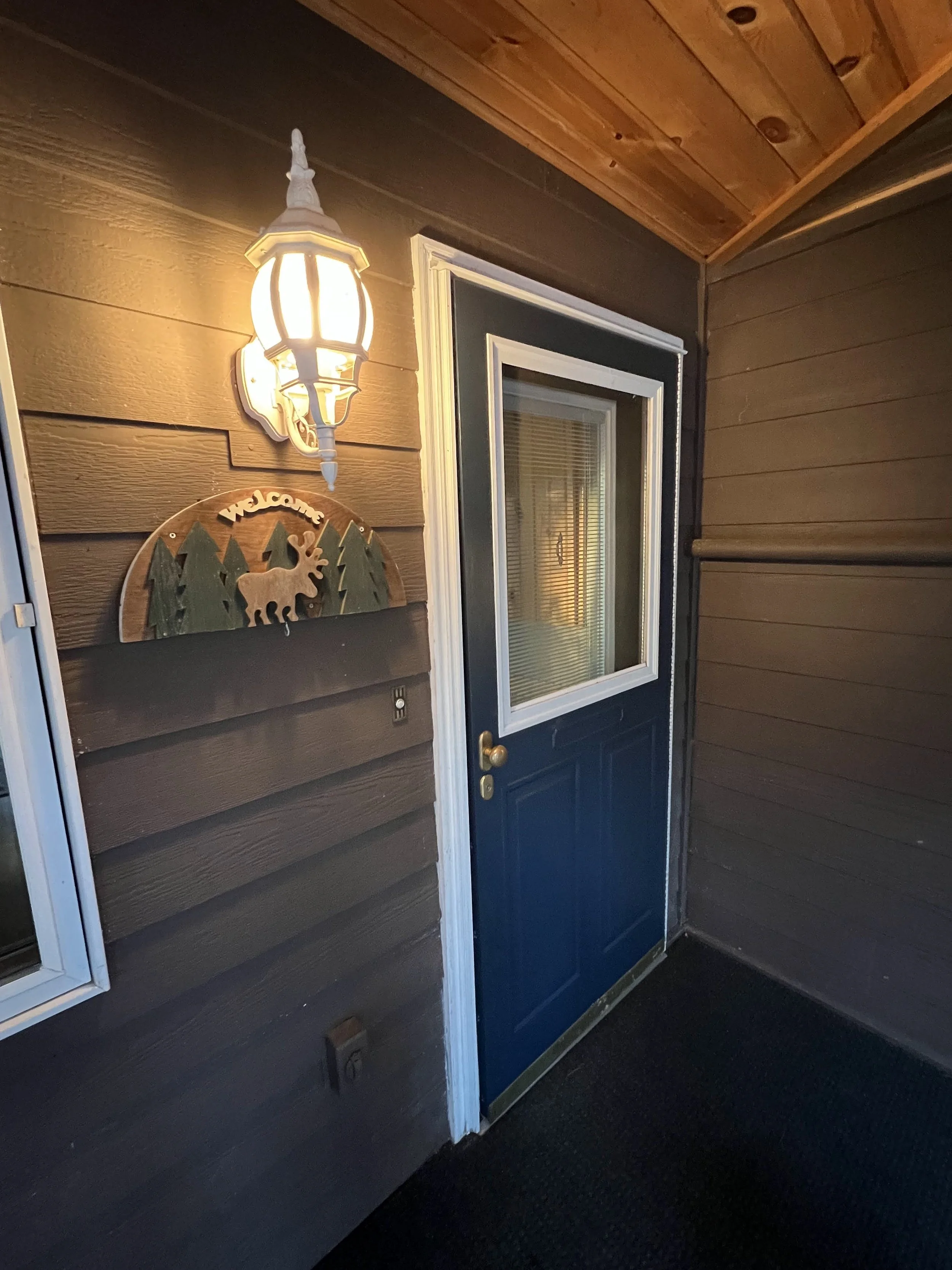 Nighttime view of a covered porch with a blue door, white trim, a window, a welcome sign with trees and a moose, and an exterior lantern light.