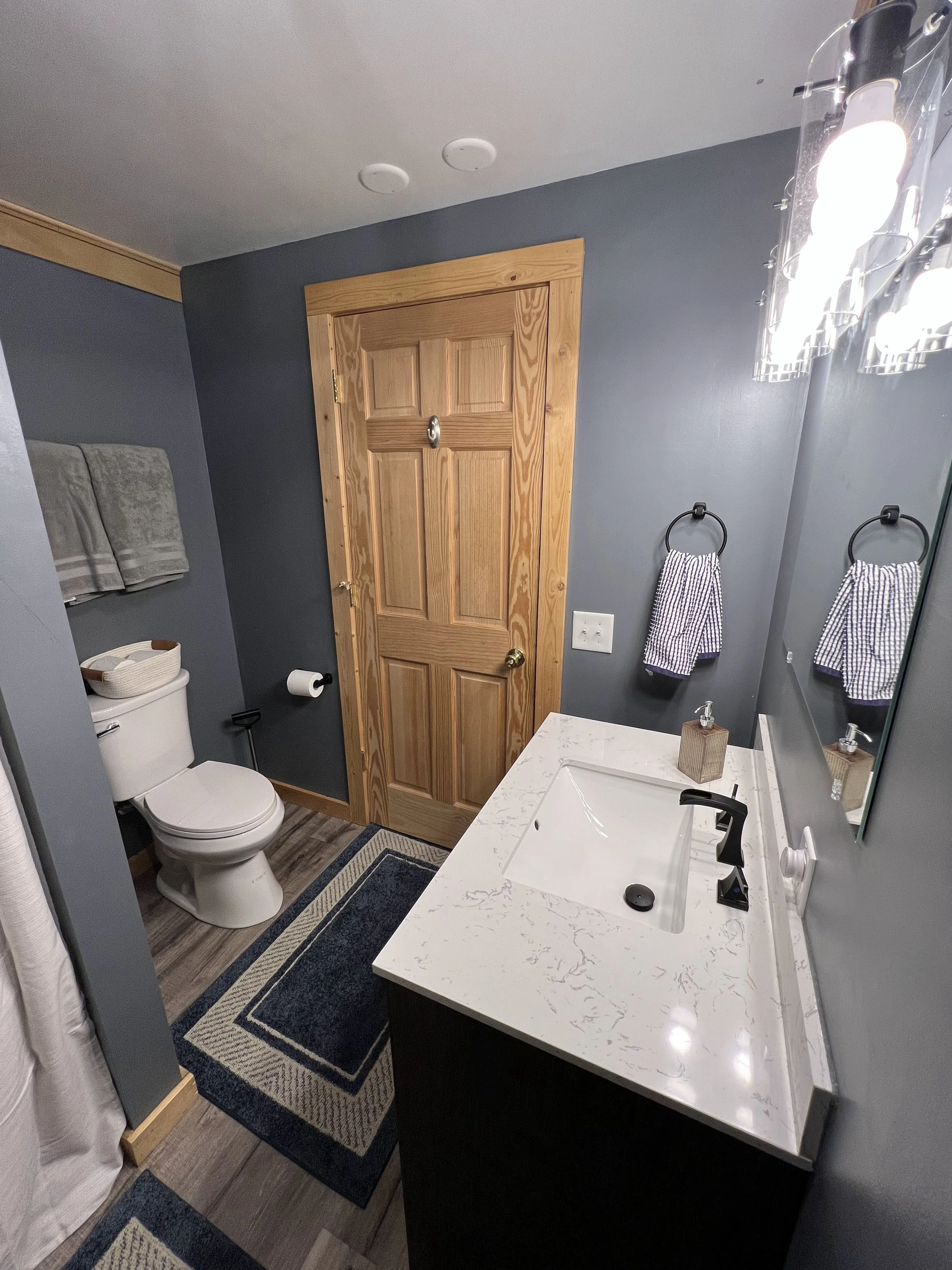 Bathroom with wooden door, gray walls, white marble countertop sink, mirror, black faucet, blue and beige rug, toilet with towel rack, towels, soap dispenser, towel rings, and ceiling lights.