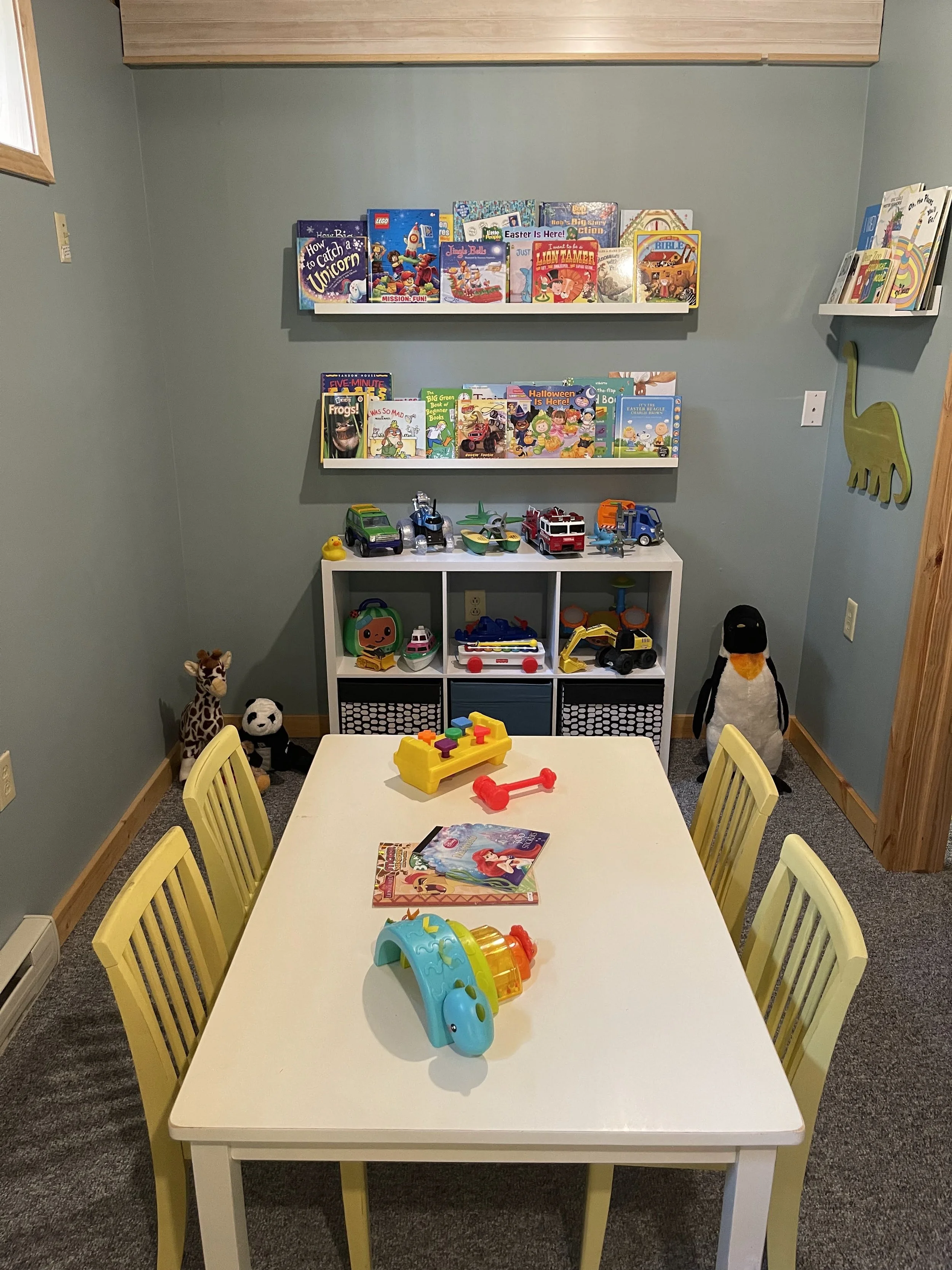 A children's playroom with a white table and yellow chairs, toys, stuffed animals, and books on shelves on the wall.