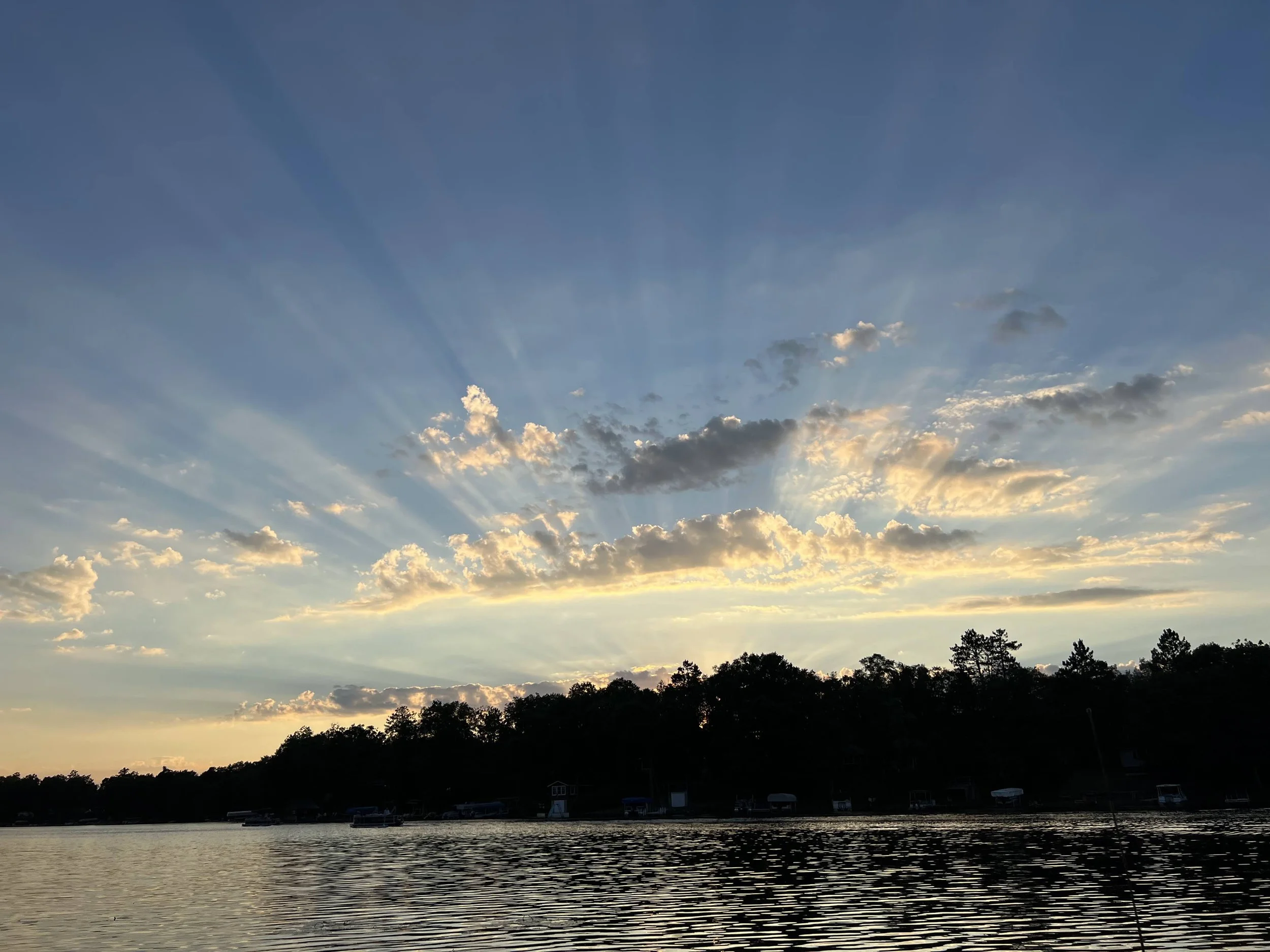 Sunset over a lake with partly cloudy sky, silhouetted trees along the shoreline, and boats docked near houses.