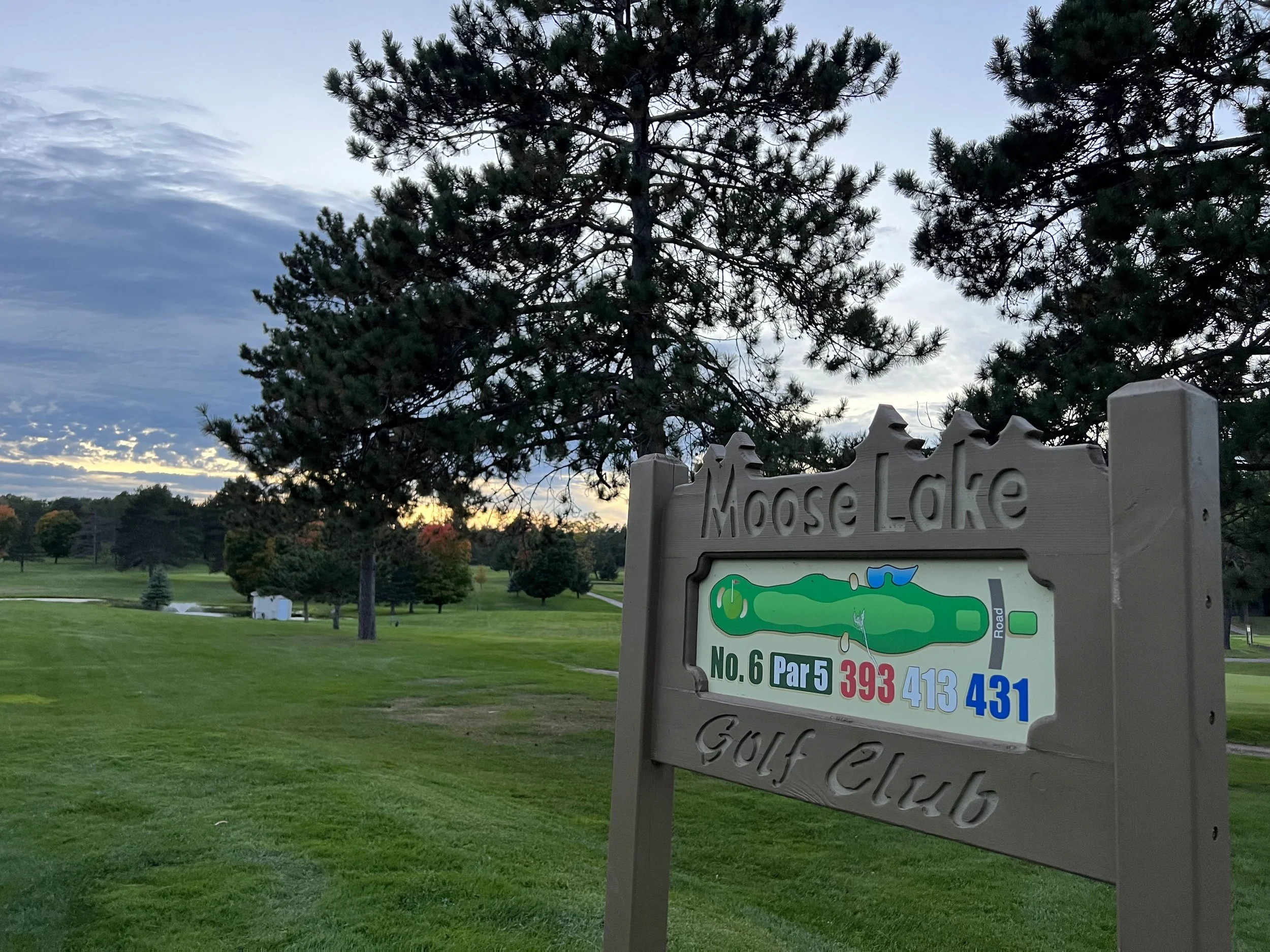 Sign at Moose Lake Golf Club showing hole number 6, a par 5, with distances 393, 413, and 431 yards, surrounded by trees and a lush green golf course at sunset.