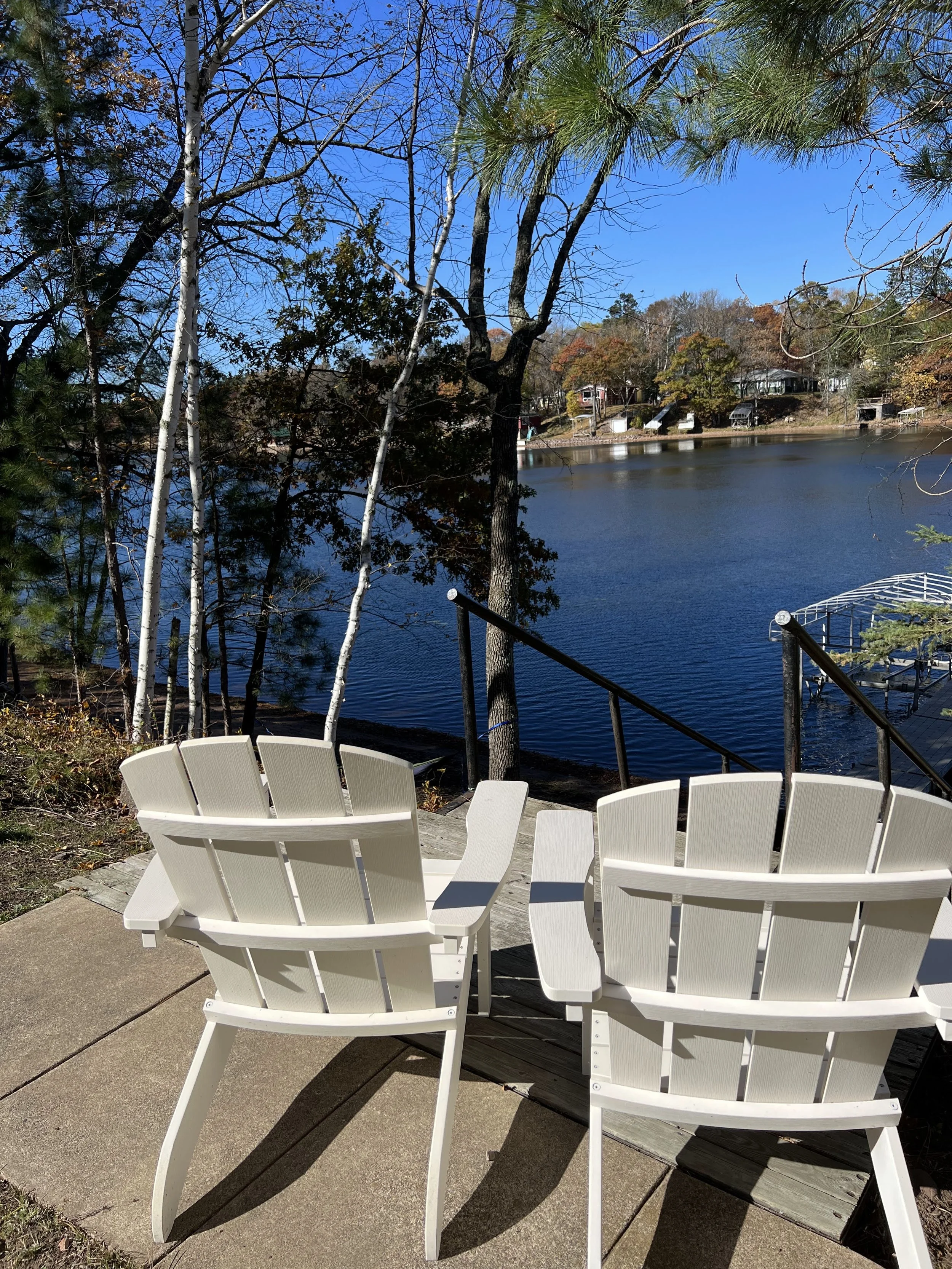 Two white Adirondack chairs on a patio overlooking a lake with trees and houses in the distance.