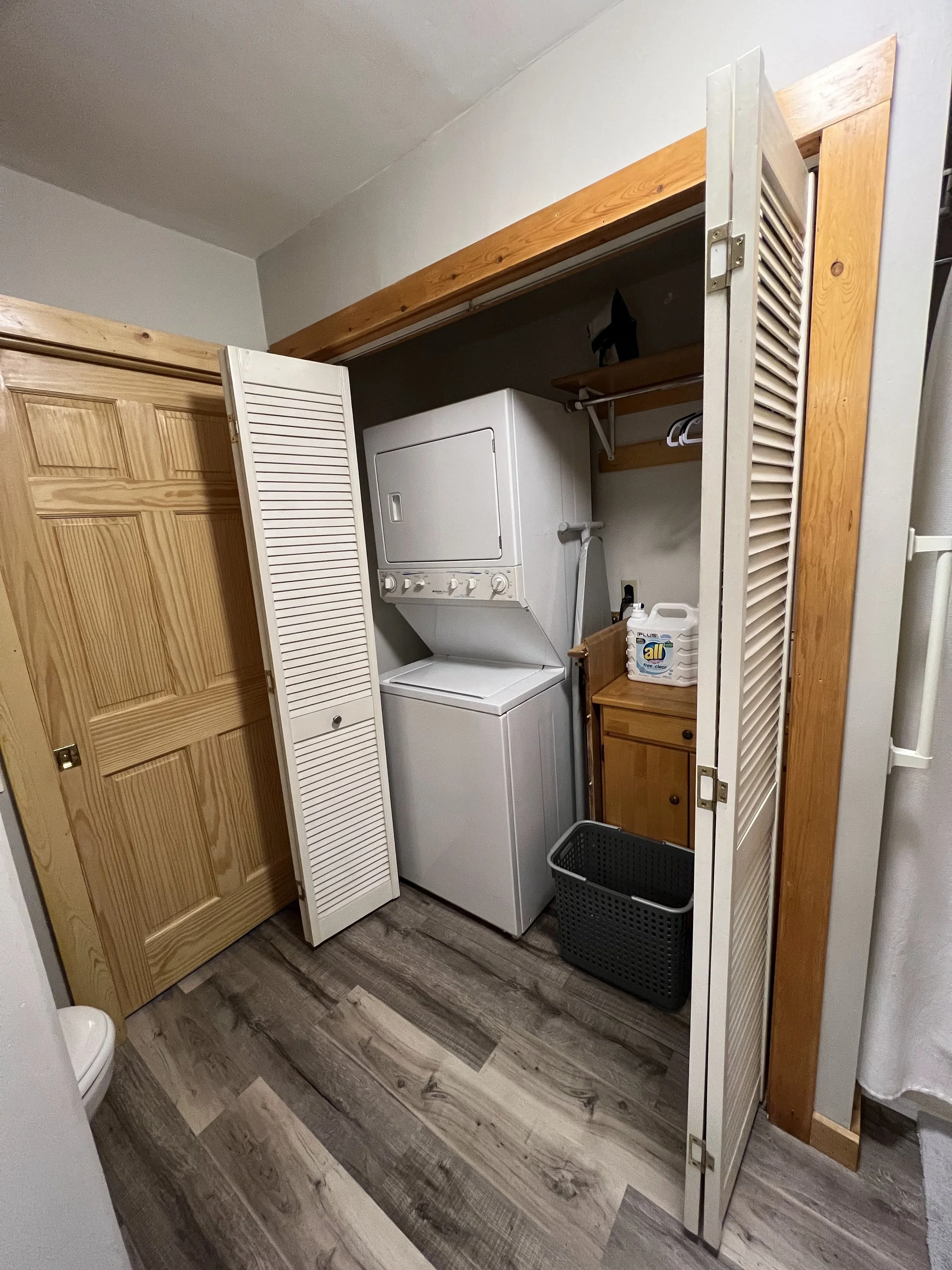 A laundry area with a stacked washer and dryer enclosed in a closet with louvered doors, a small wooden cabinet, a laundry basket, and shelves for storage.