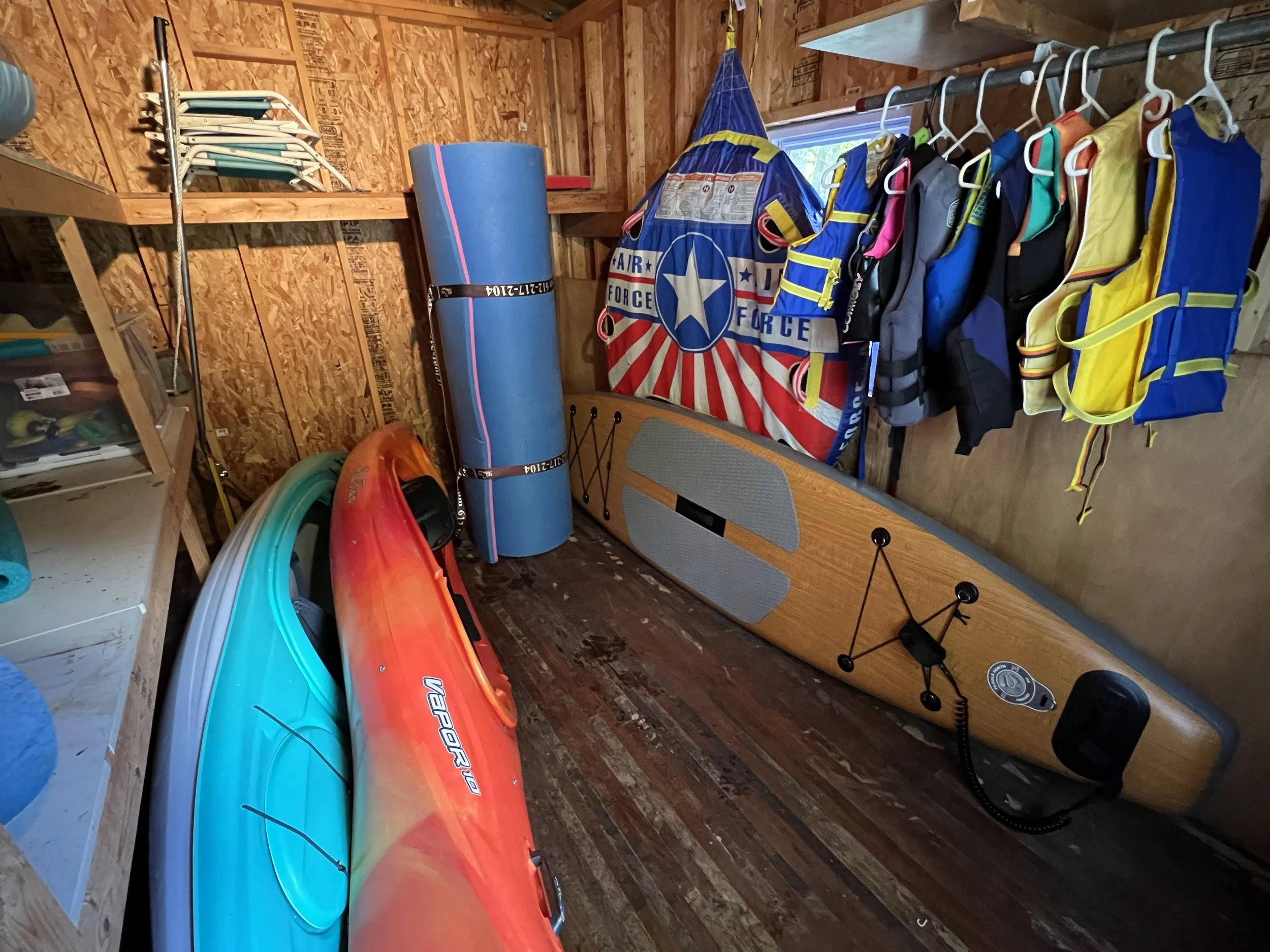 A garage storage space with colorful kayaks, a paddleboard, and life jackets hanging on the wall.