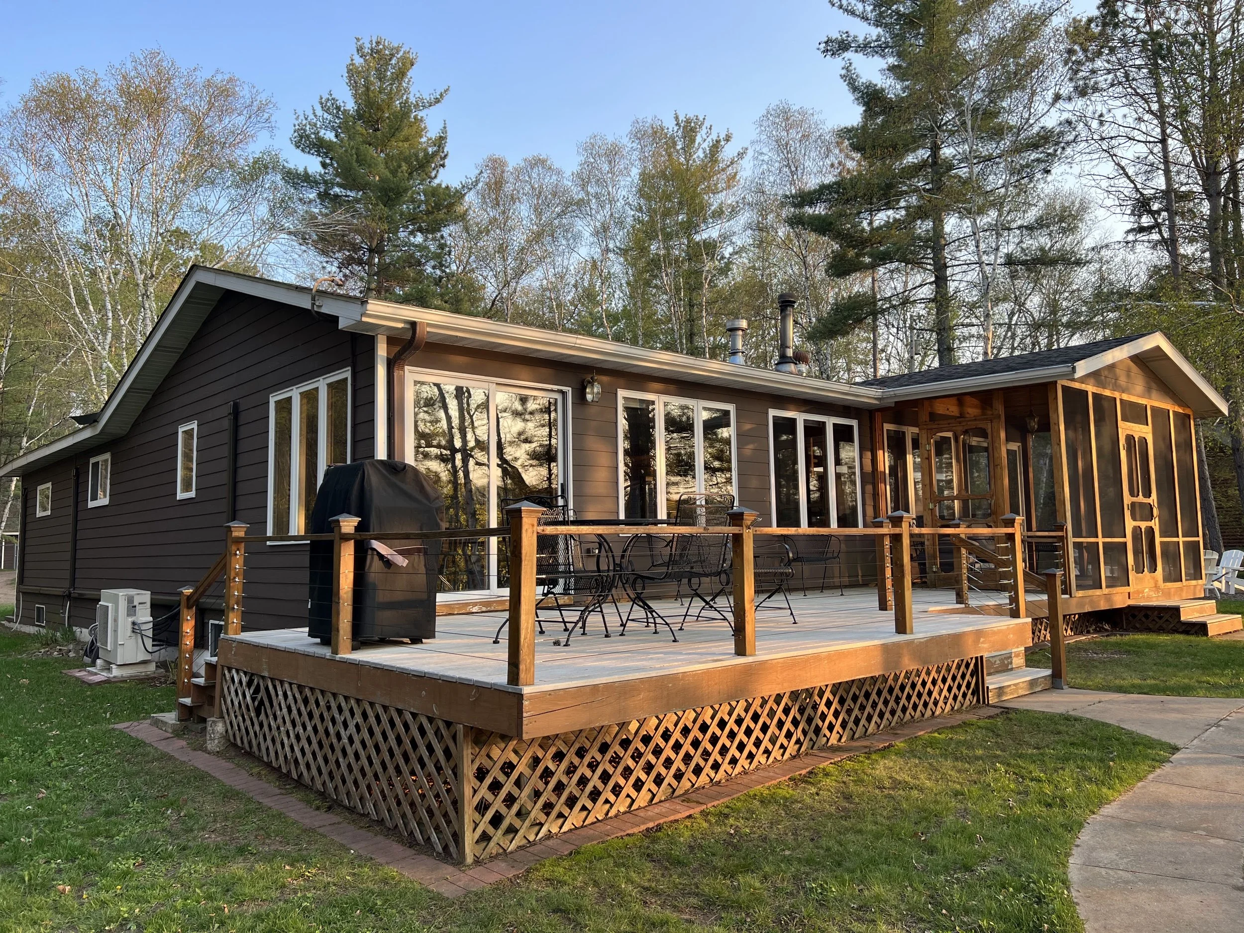A house with a large wooden deck, black siding, multiple sliding glass doors, and surrounded by tall trees during sunset.