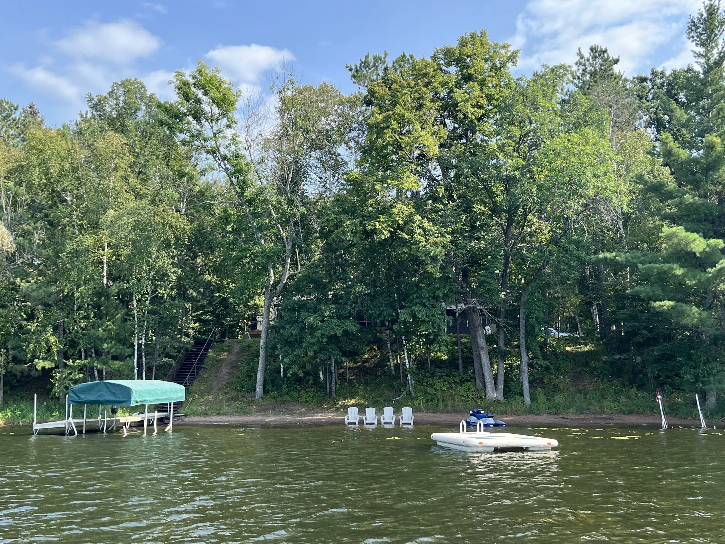 Lakeside scene with a dock and boat cover, four white Adirondack chairs on the shore, and lush green trees under a partly cloudy sky.