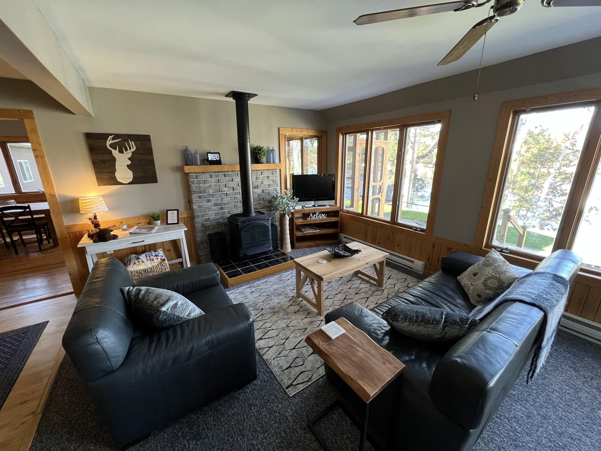 Living room with two black leather sofas, a wooden coffee table, a wood-burning stove with a brick surround, large windows with wood trim, a TV on a wooden stand, a white side table with a lamp, and decorative items, in a cozy cabin-style interior.