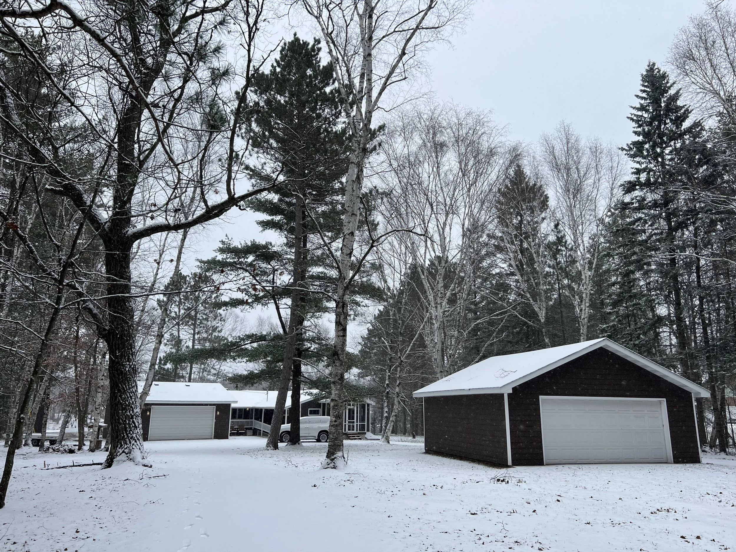 A snowy backyard with tall trees, a dark brown garage with a white door, and a house with a porch in the background.