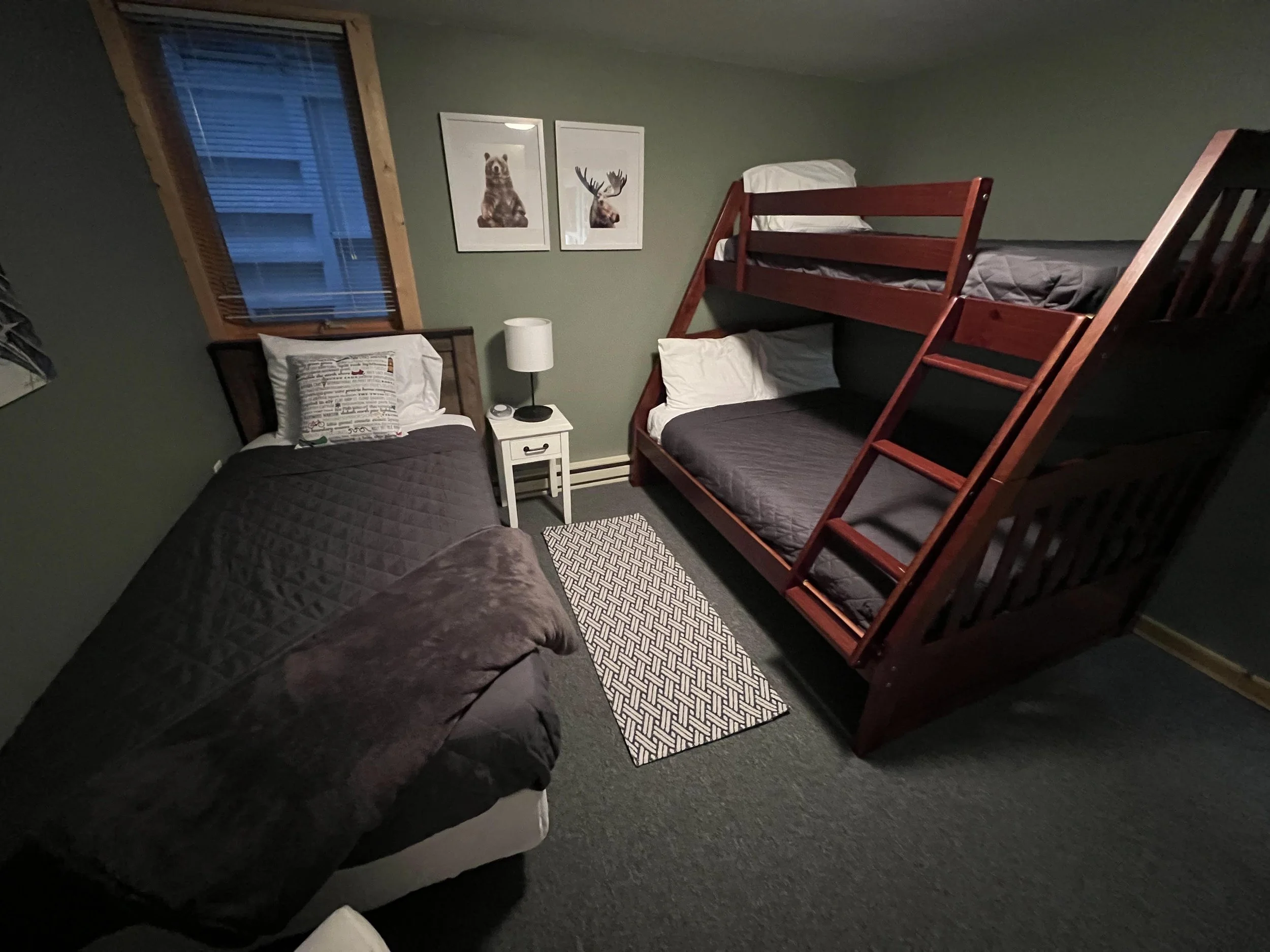 A bedroom with green walls, featuring a twin bed with black bedding and a patterned pillow, a white nightstand with a lamp and clock, two framed animal artwork (bear and moose), a window with wooden frame and blinds, and a wooden bunk bed with gray b