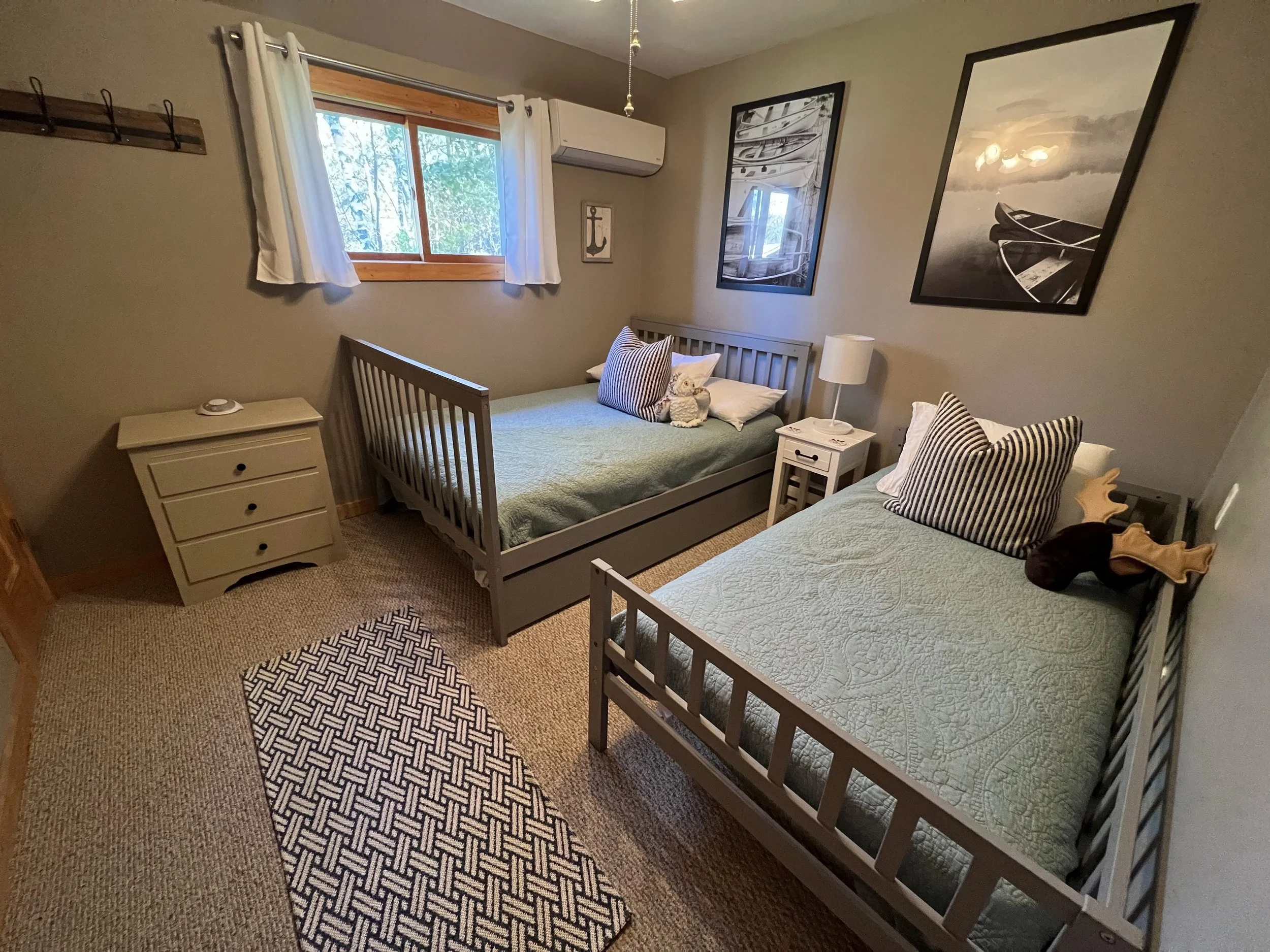 A cozy bedroom with two beds, one on each side, decorated with nautical-themed artwork, a window with white curtains, a small nightstand with a lamp, a dresser, and a patterned rug on beige carpet.