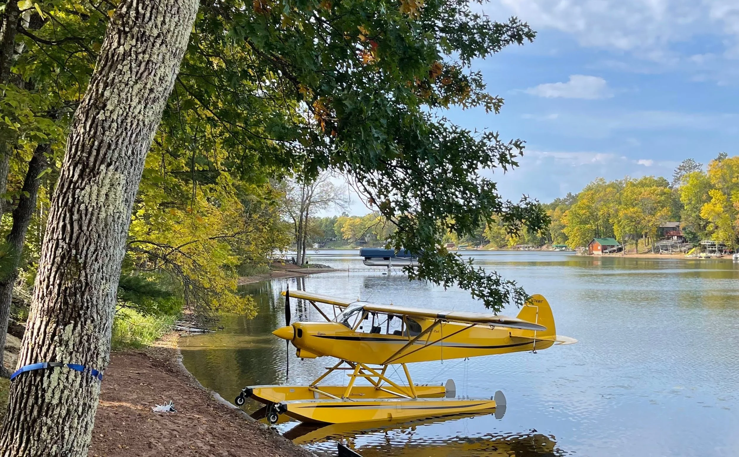 A yellow yellow amphibious airplane docked at the shore of a peaceful lake, surrounded by green trees and a clear blue sky.
