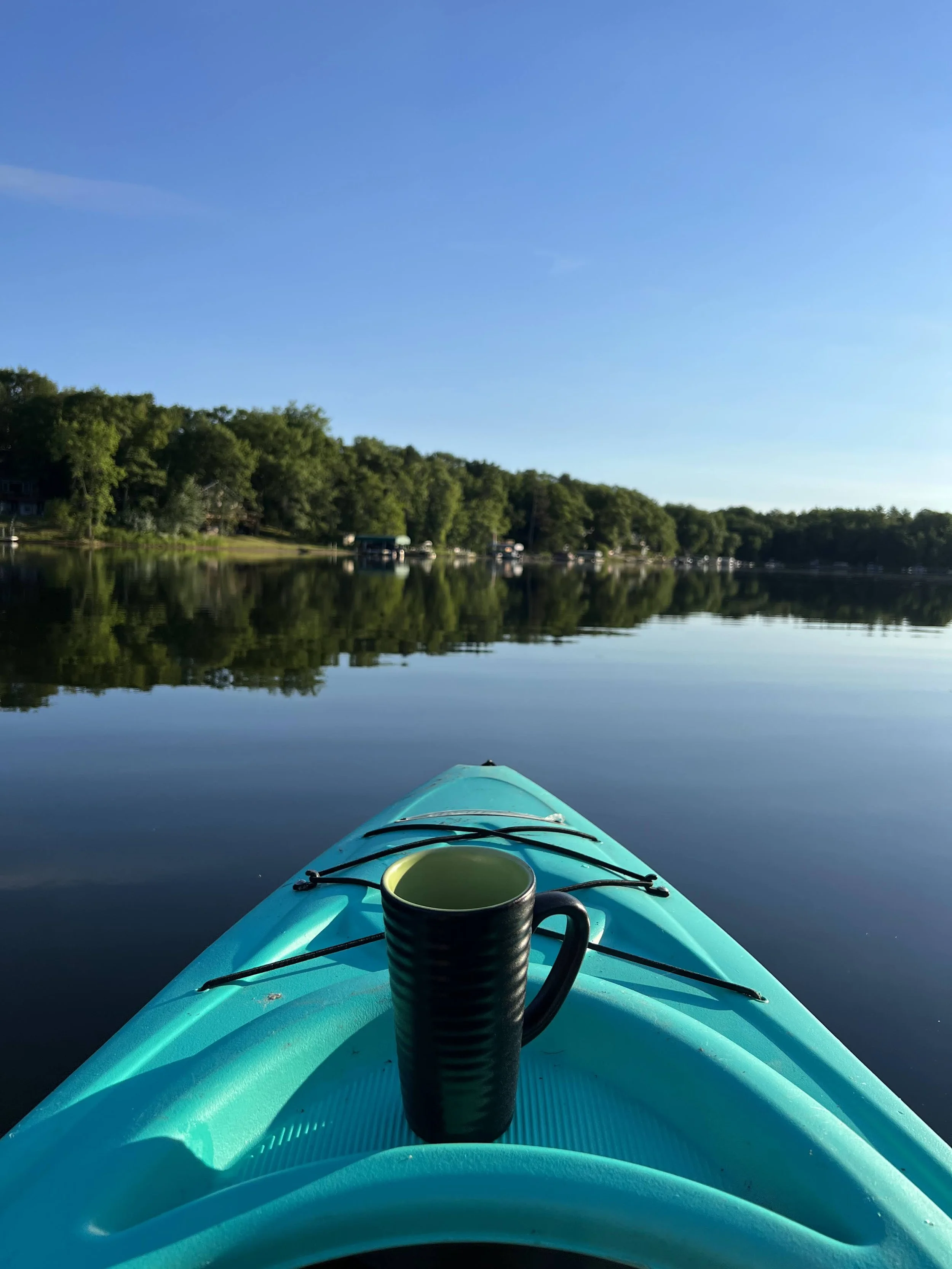 Kayak on a calm lake with a black coffee mug placed on the front, surrounded by trees and a clear blue sky.