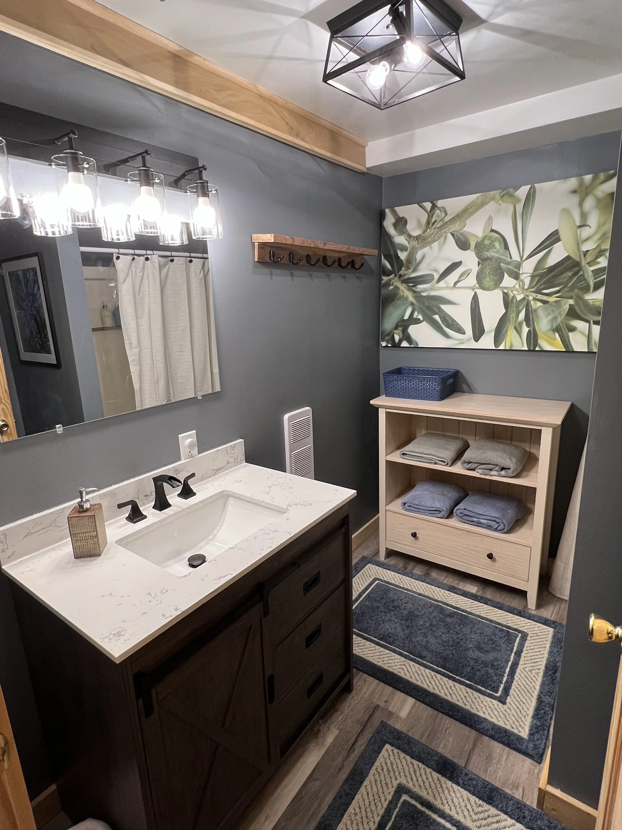 A bathroom with a dark wooden vanity, marble countertop, and black fixtures. There is a large mirror, a wall-mounted light fixture with multiple bulbs, a wooden shelf with hooks, a framed photo, a large canvas of greenery, and a small wooden shelf wi